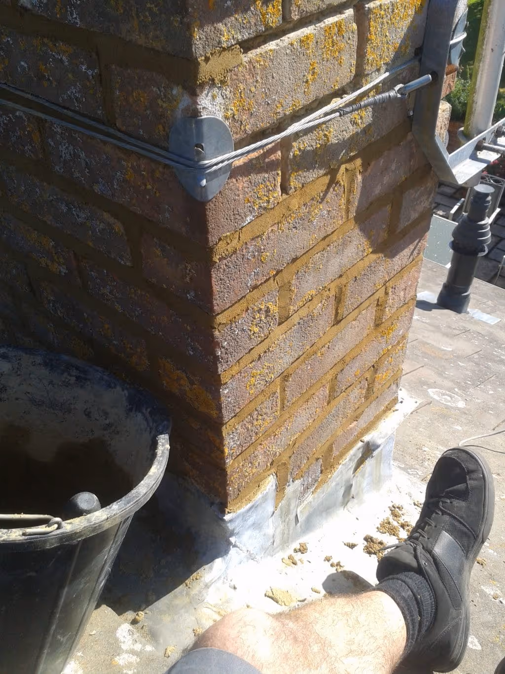 Close-up of a brick chimney corner with fresh mortar repointing, metal stays attached, and a person's leg wearing a black shoe resting nearby.