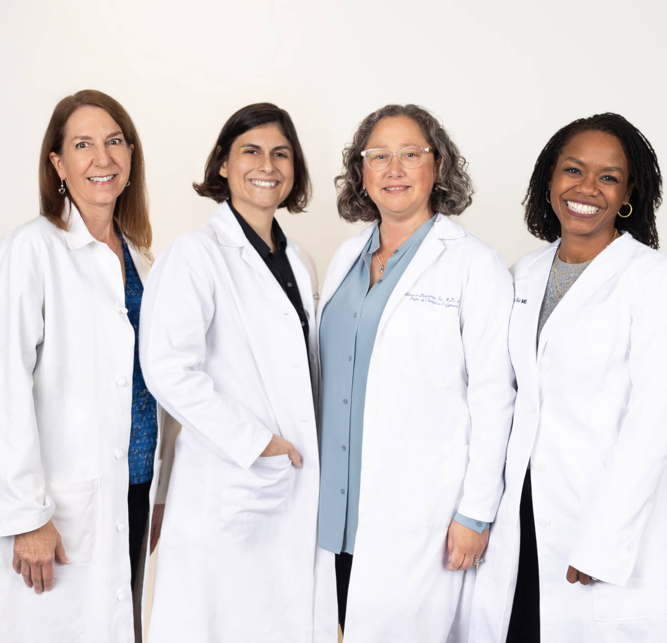 Four people stand together in white doctors coats, against a white wall, smiling at the camera.