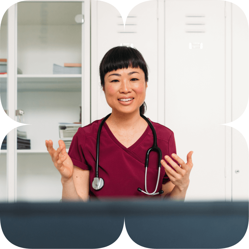 A doctor smiles at the camera, standing in front of a desk, while using their hands to explain while they talk.
