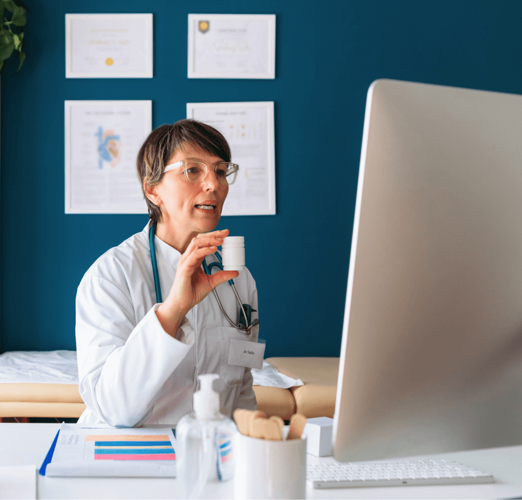 A doctor sitting in a blue room shows a medication during a video call.
