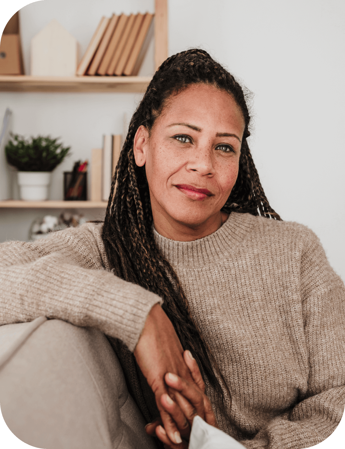 A person in a beige sweater looks peacefully at the camera while sitting in a warm room with white walls and an inviting couch. 