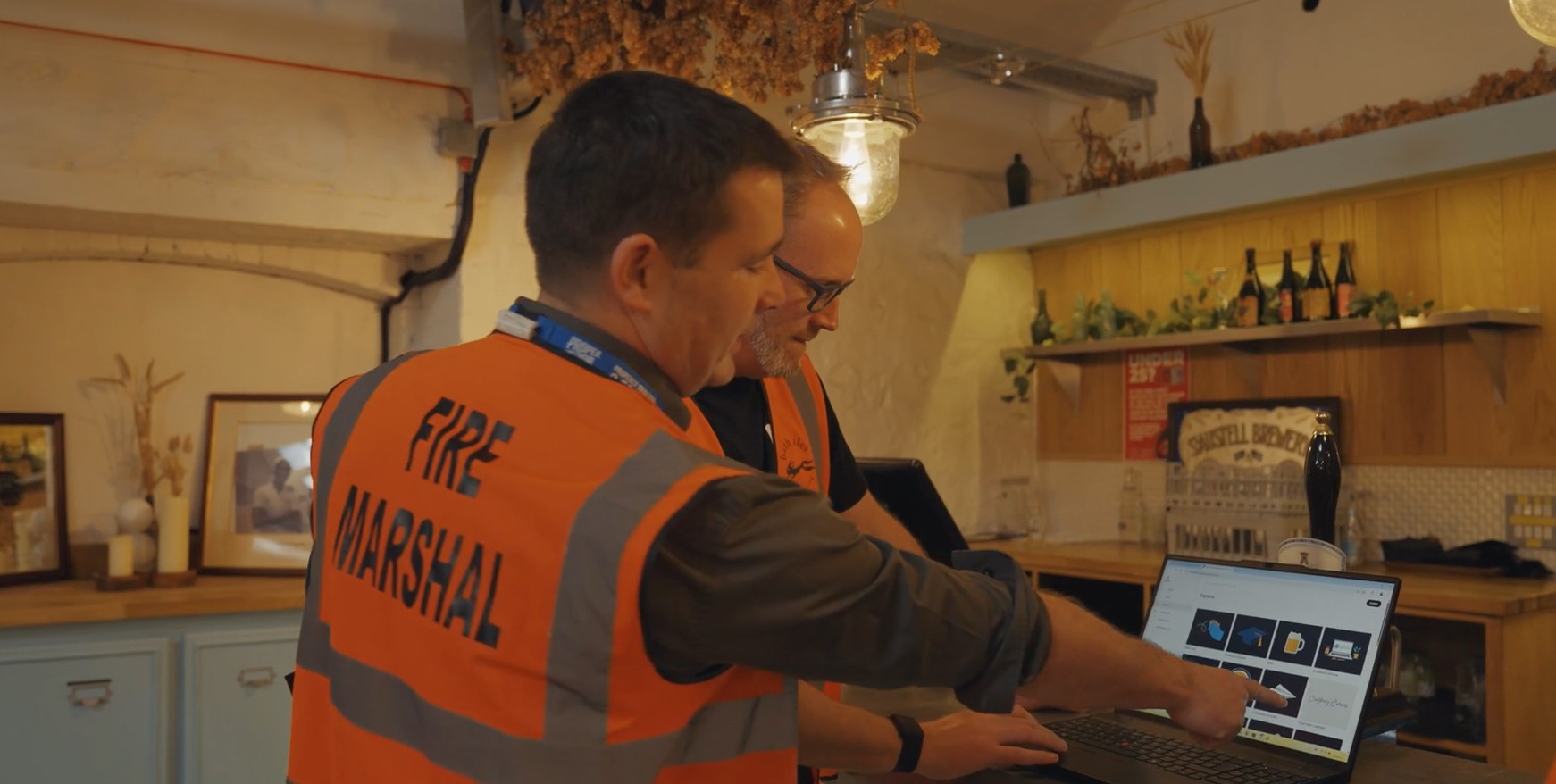 St Austell employee wearing high-vis FIRE MARSHALL vest looking at compliance training on laptop, accompanied by Thrive employee also in a high-vis vest