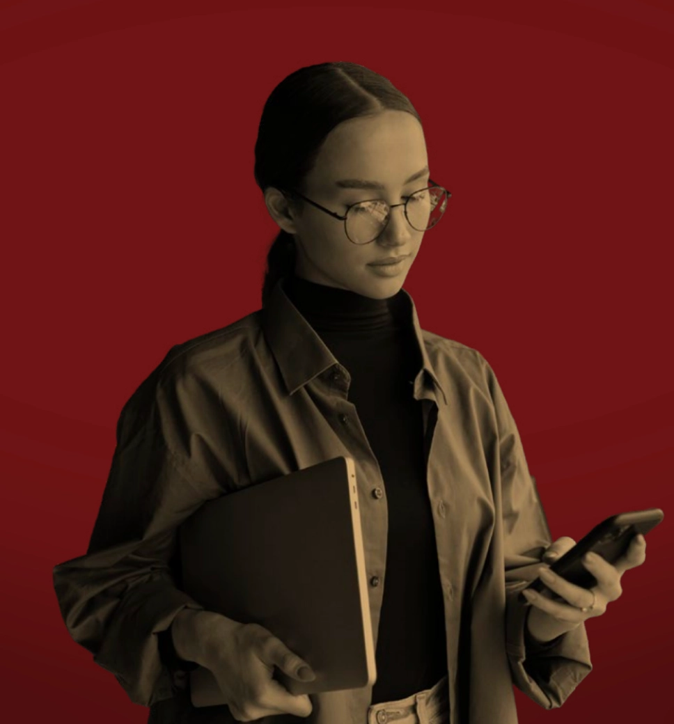 A sepia-toned person wearing glasses and holding a laptop looks down at a smartphone against a solid red background.