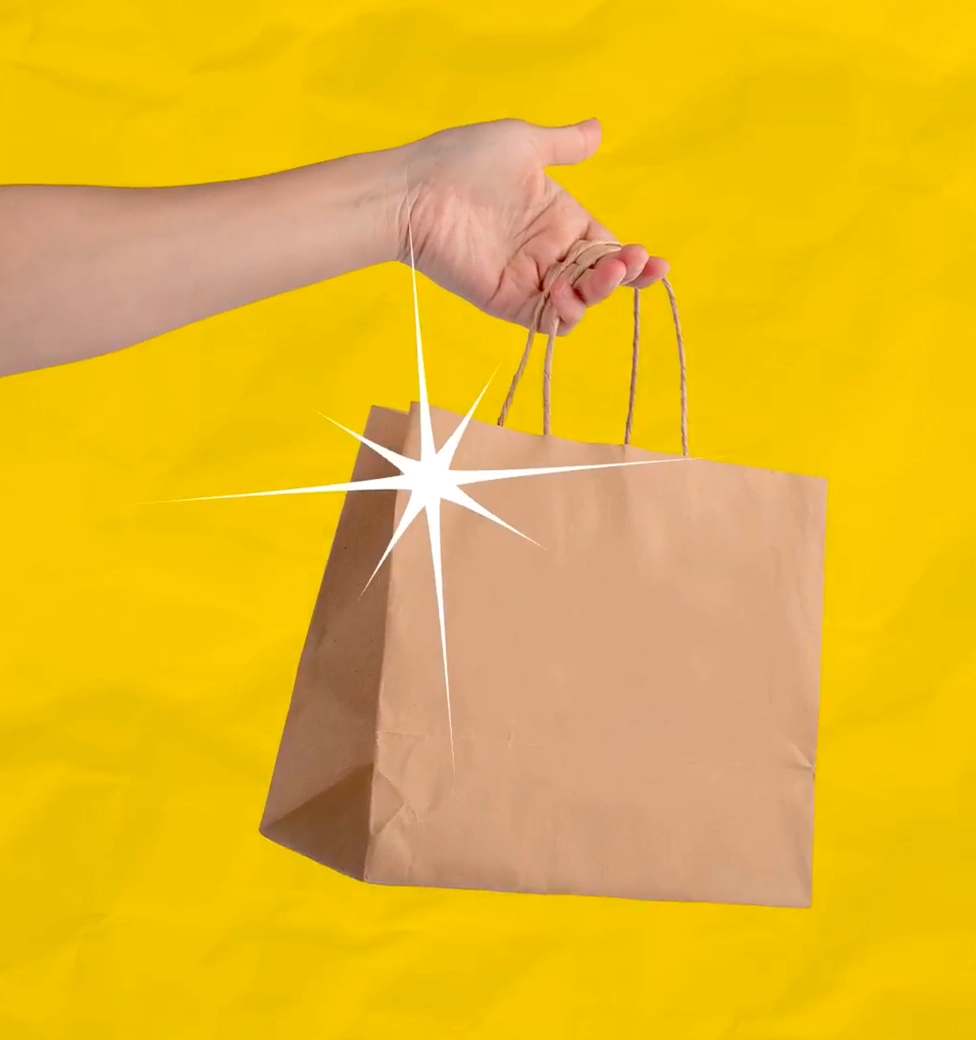 A hand holds a plain brown paper shopping bag against a bright yellow background, with a white sparkle graphic on the bag.