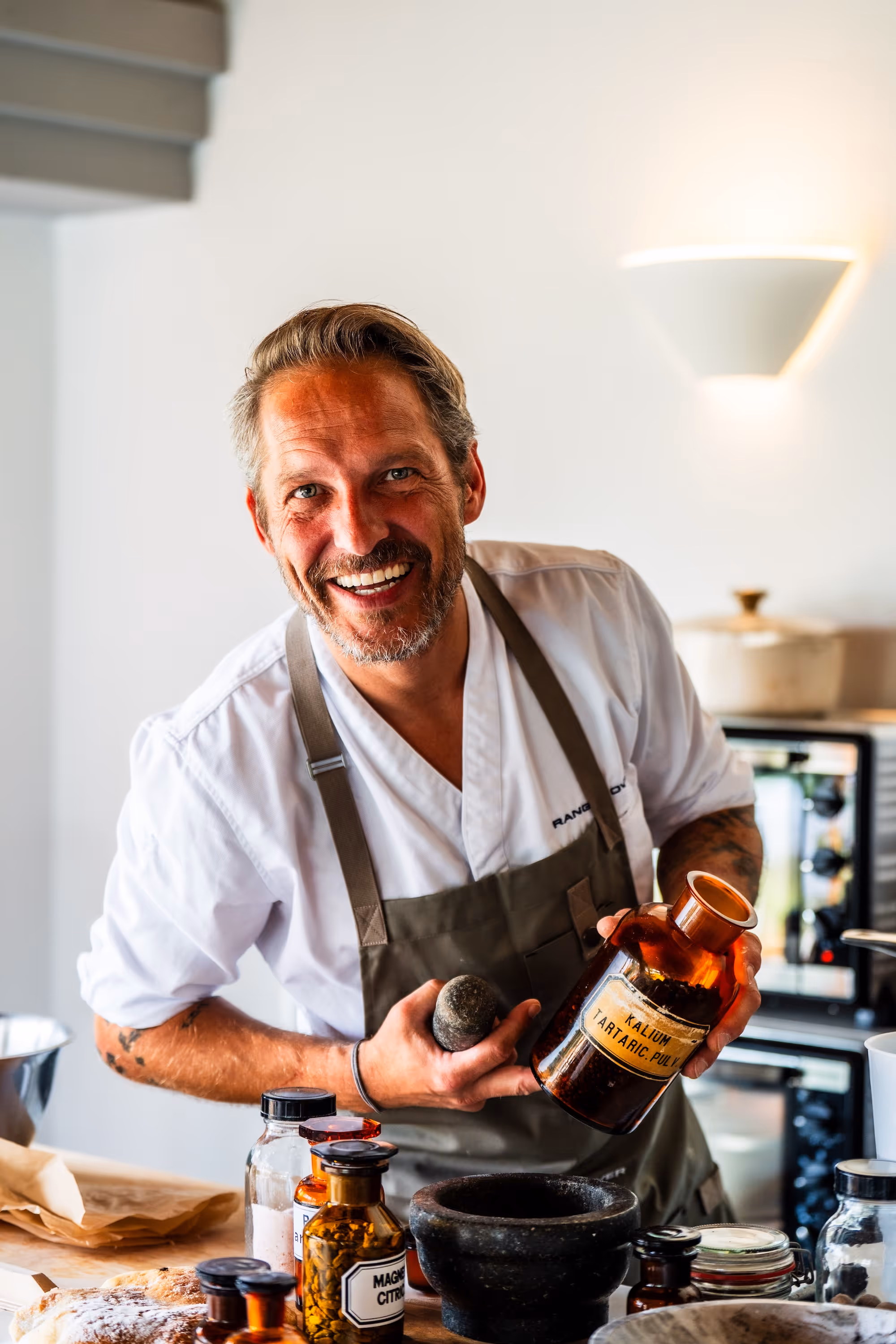 Koch Rainer Klutsch lacht herzlich in die Kamera. Er hält eine braune Apothekerflasche und einen Stößel in der Hand, vor ihm auf dem Tisch stehen Mörser und weitere Gewürze.