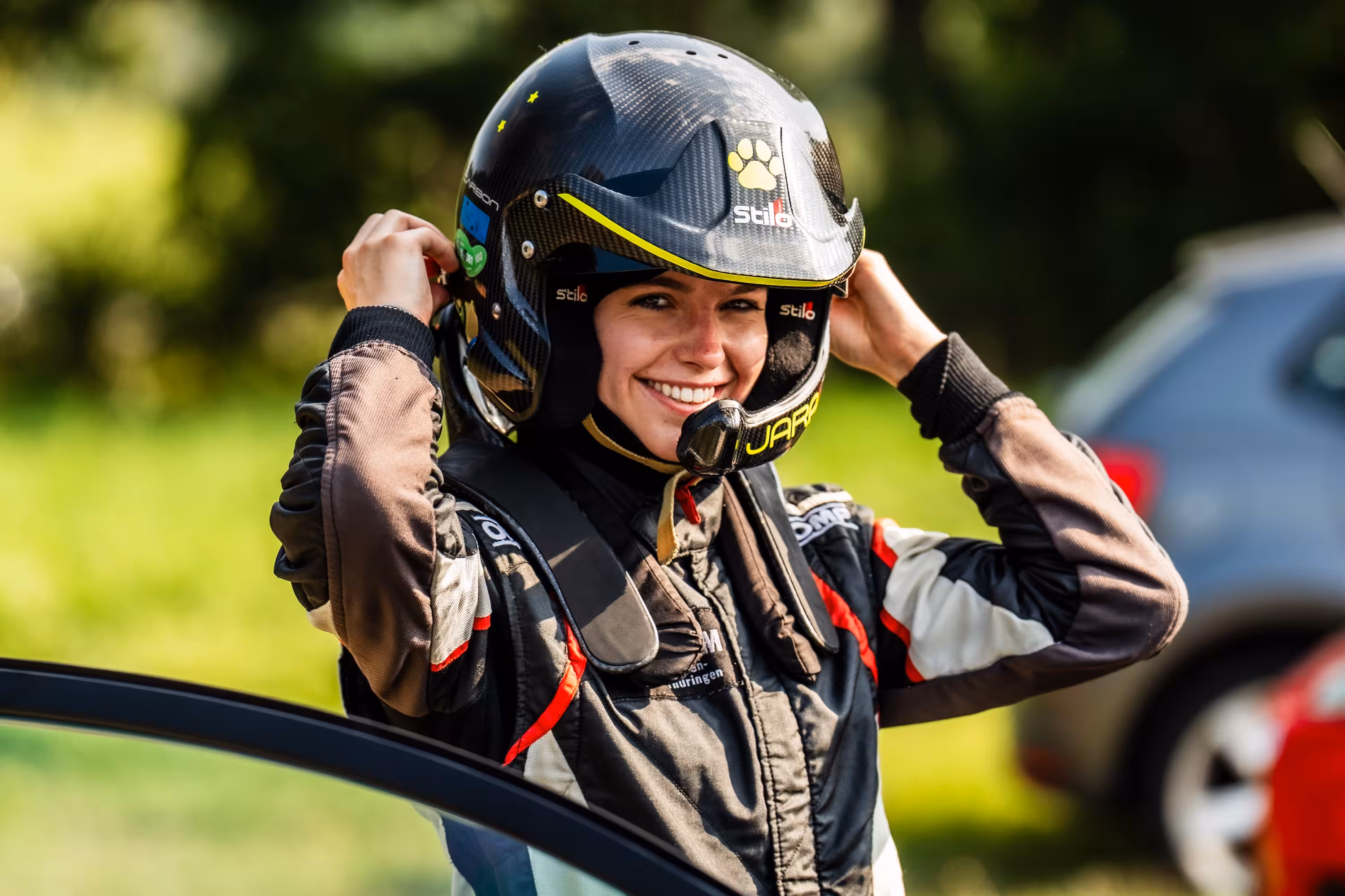 Jara Hain, eine Rallye-Beifahrerin, lacht in die Kamera, während sie sich ihren schwarzen Stilo-Helm mit gelben Akzenten aufsetzt. Sie trägt einen Rennanzug mit den Farben Schwarz, Weiß und Rot.