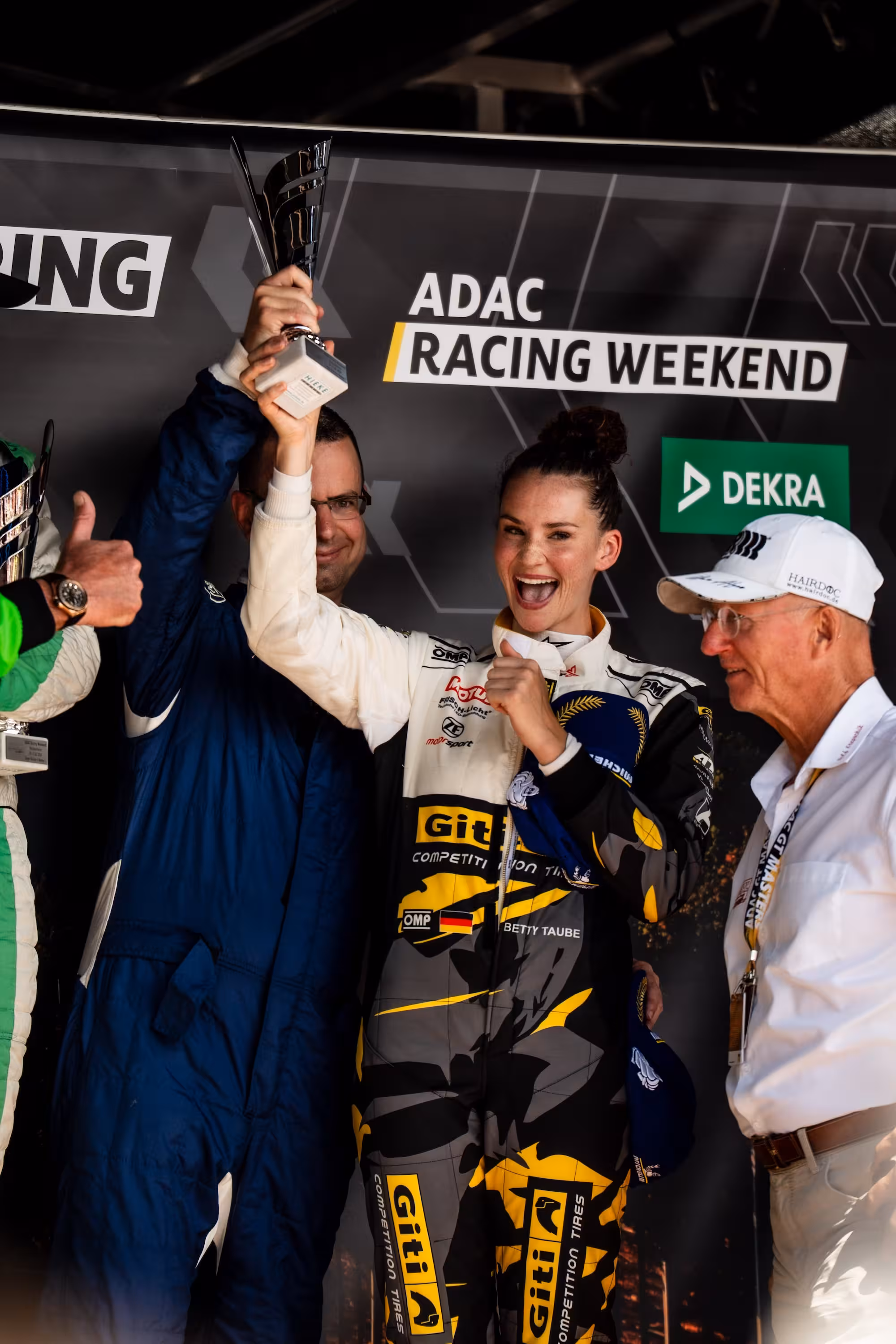 Model, Influencerin und Rennfahrerin Betty Taube jubelt auf dem Podium des ADAC Racing Weekends. Sie trägt einen Rennanzug im Giti-Design und hält stolz ihren silbernen Pokal in die Höhe, unterstützt von einem Teammitglied.