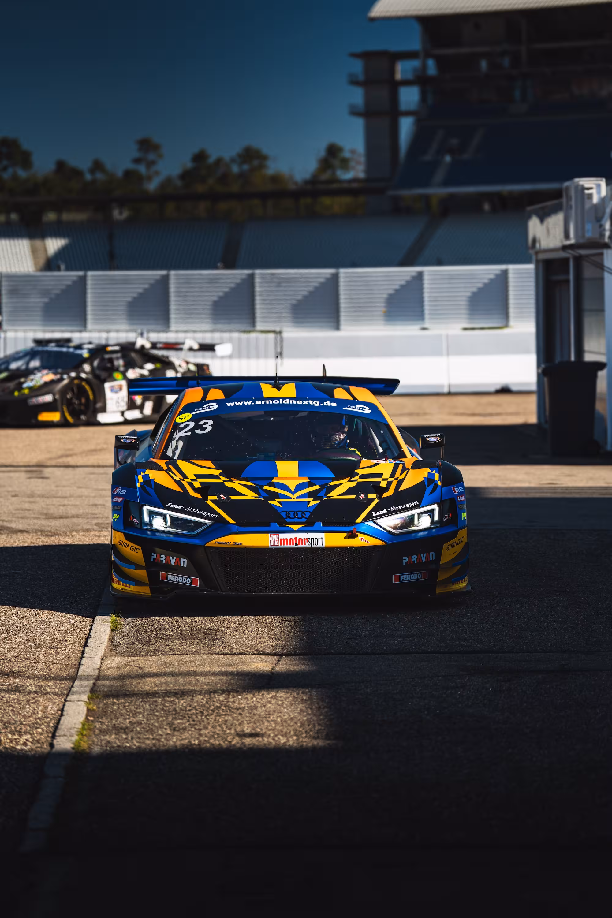 Frontansicht eines blau-gelben Audi R8 LMS in der Boxengasse des Hockenheimrings. Das Fahrzeug steht im harten Sonnenlicht vor den Tribünen beim ADAC Racing Weekend 2024.