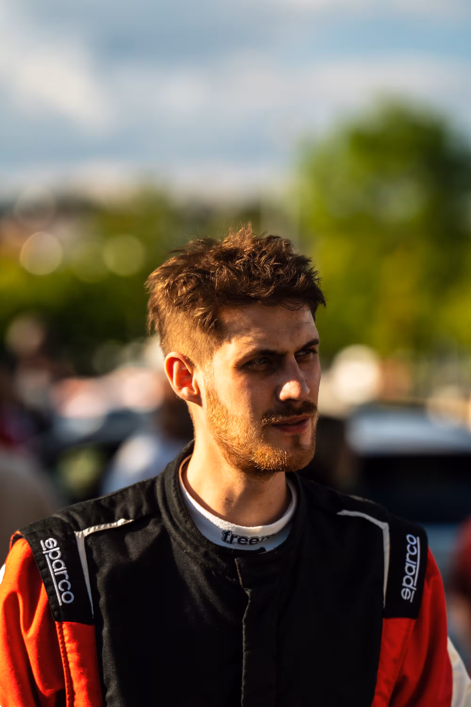 Porträt des Rallyefahrers Florian Schwenker, der mit ernstem Ausdruck zur Seite blickt. Er trägt einen schwarz-roten Rennanzug der Marke Sparco.