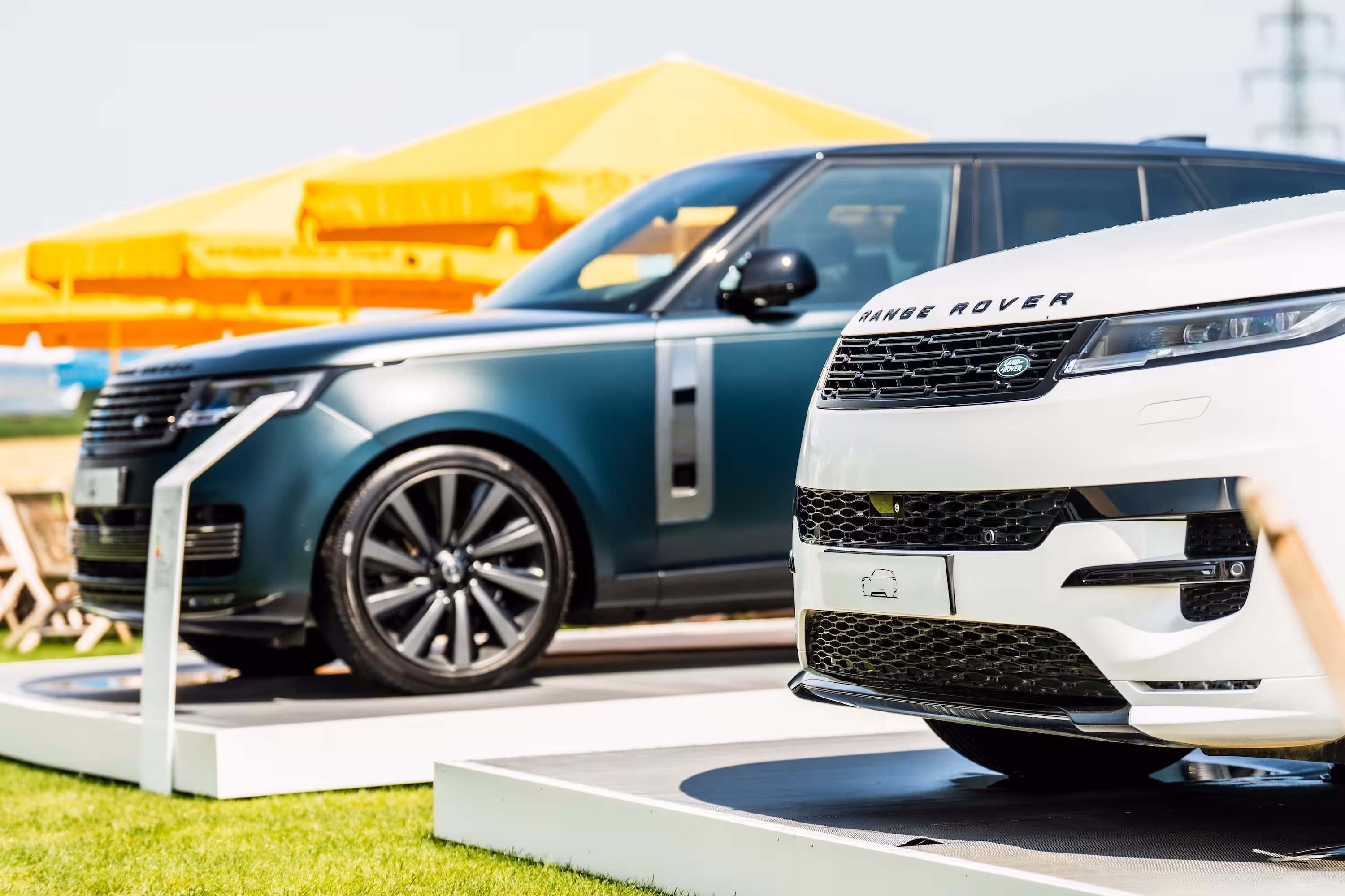 Nahaufnahme der Front eines weißen Range Rover Sport, der auf einem weißen Podest steht. Im Hintergrund ist unscharf ein dunkelgrüner Range Rover vor gelben Zelten zu sehen. Die Szene ist Teil der Ausstellung bei der German Polo Tour in München.