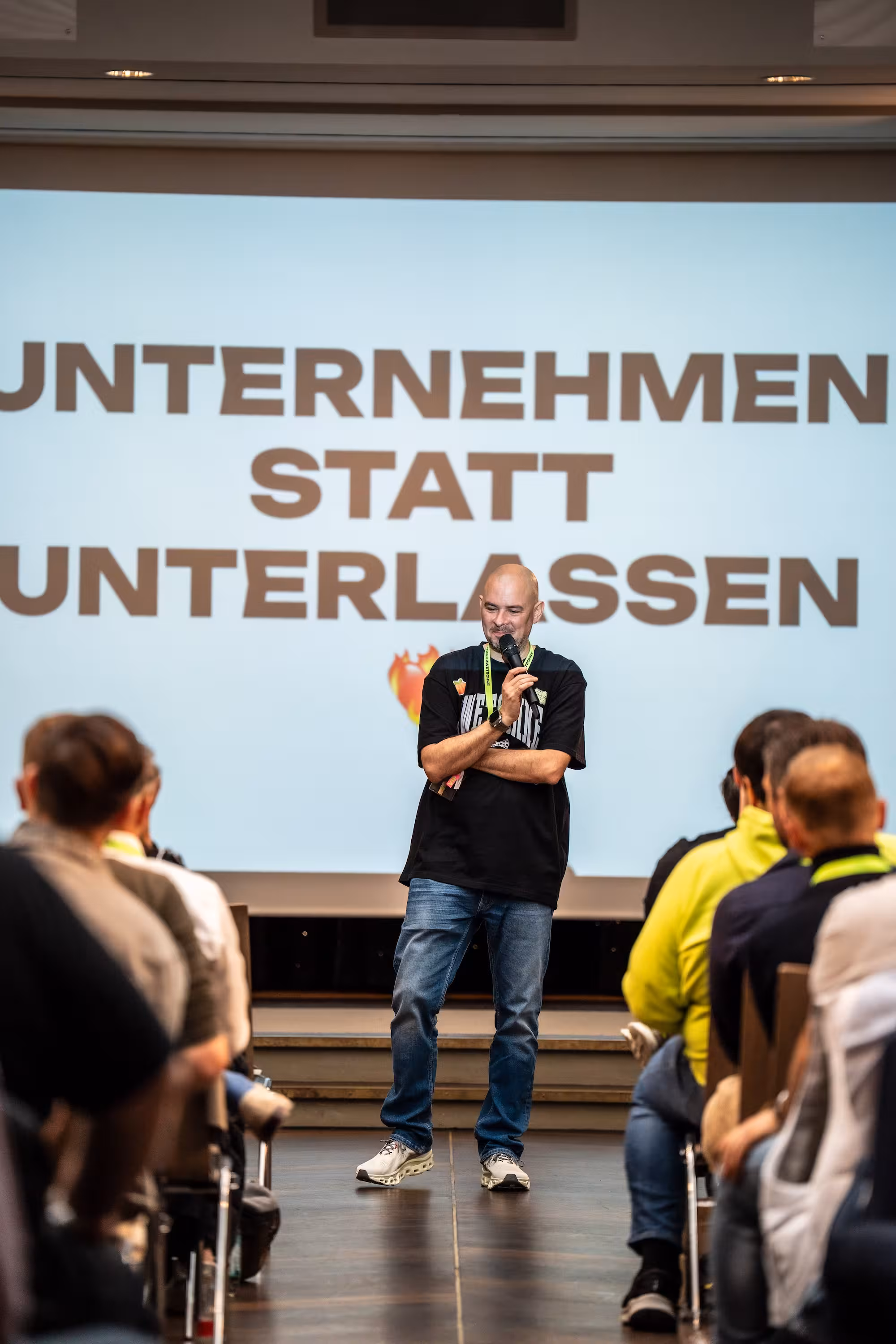 Hannes Zwetschke steht lachend auf der Bühne des Hotel Maximilian's. Hinter ihm leuchtet der Slogan 'UNTERNEHMEN STATT UNTERLASSEN'. Er hält ein Mikrofon und spricht zum Publikum.
