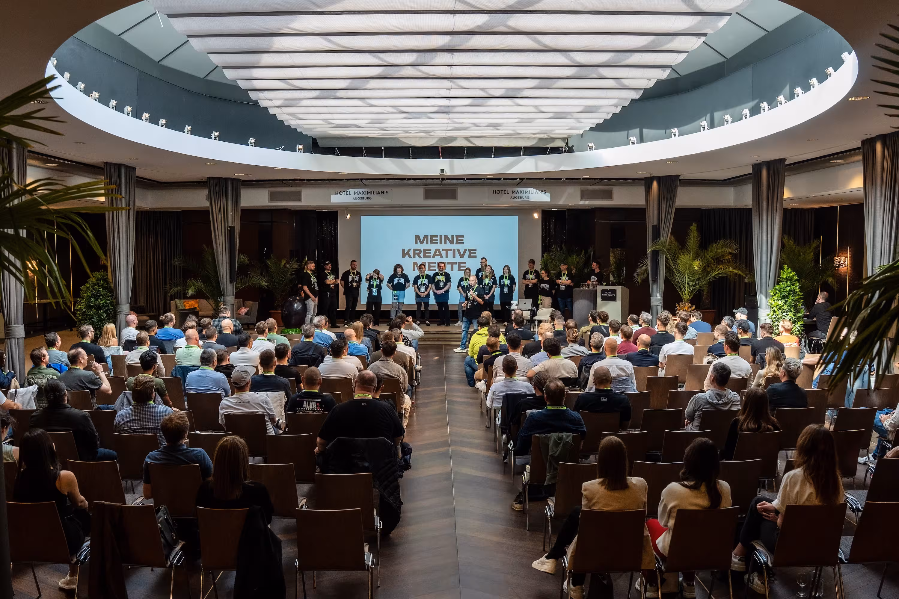Weitwinkelaufnahme des Saals im Hotel Maximilian's. Hannes Zwetschke steht mit seinem gesamten Team auf der Bühne vor der Leinwand mit der Aufschrift 'MEINE KREATIVE MEUTE'. Der Saal ist voll besetzt.