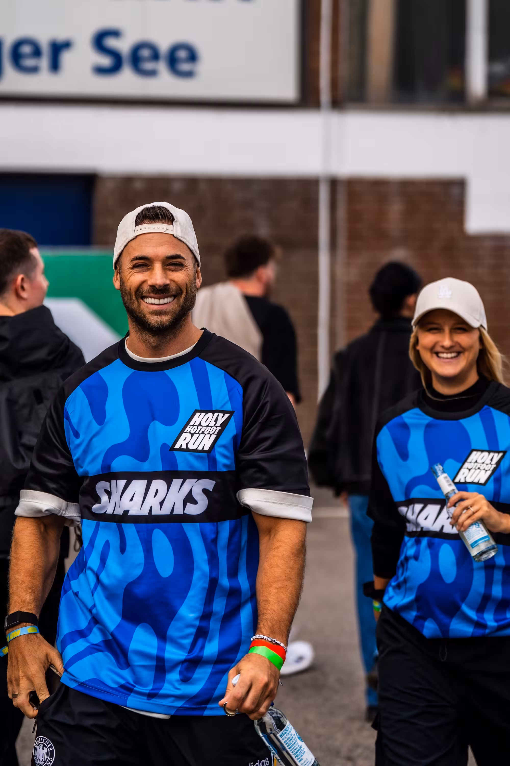 Die beiden Stuttgarter Influencer Benny & Angie, Teamchefs des Teams 'Sharks' in blau-schwarzen Trikots laufen lächelnd über den Asphalt. Benny im Vordergrund trägt eine weiße Kappe, Angie hält eine Flasche.