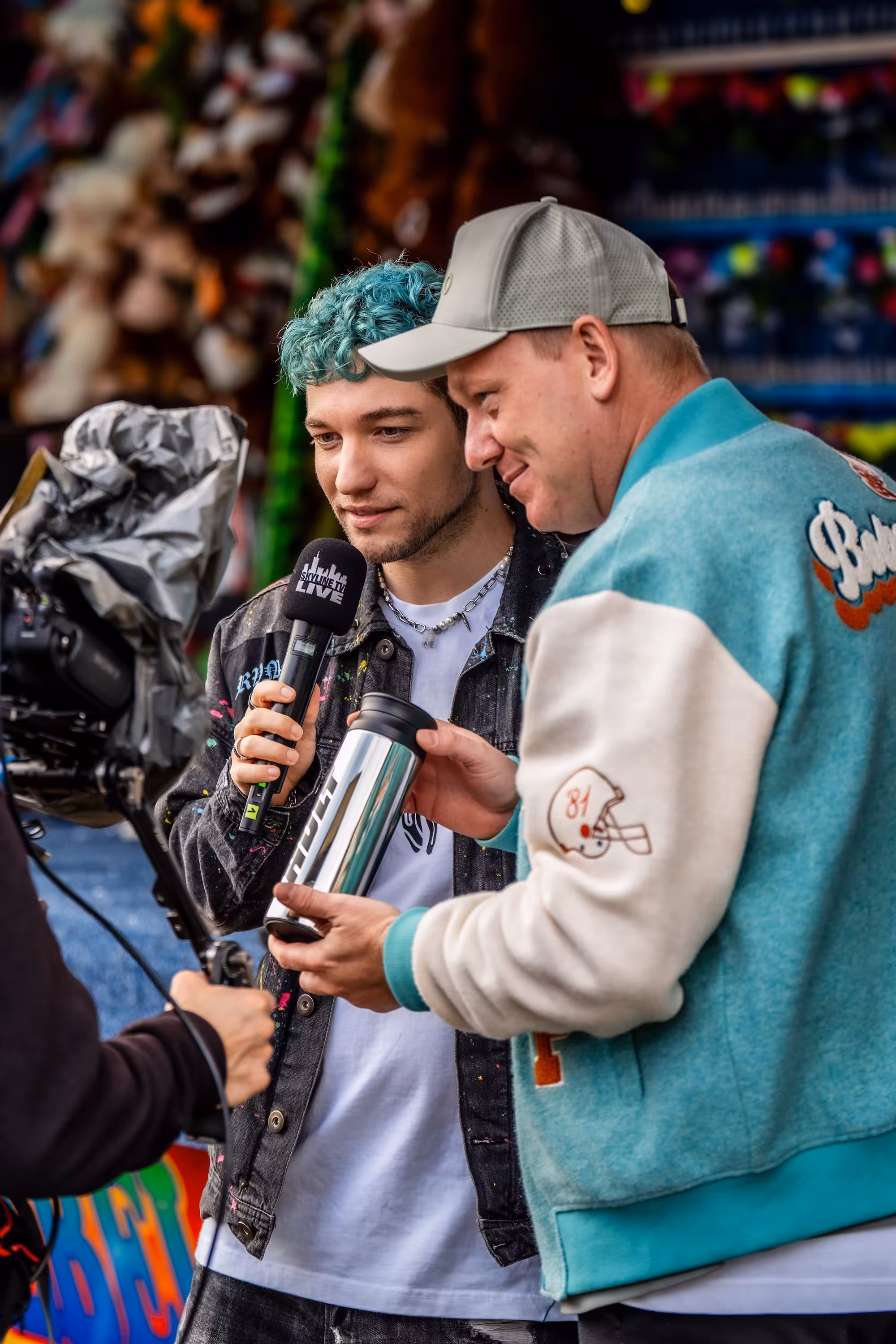 Rezo hält ein Mikrofon von 'Skyline TV Live' und steht neben Knossi. Beide schauen interessiert auf einen silbernen HOLY Thermobecher, während sie gefilmt werden.