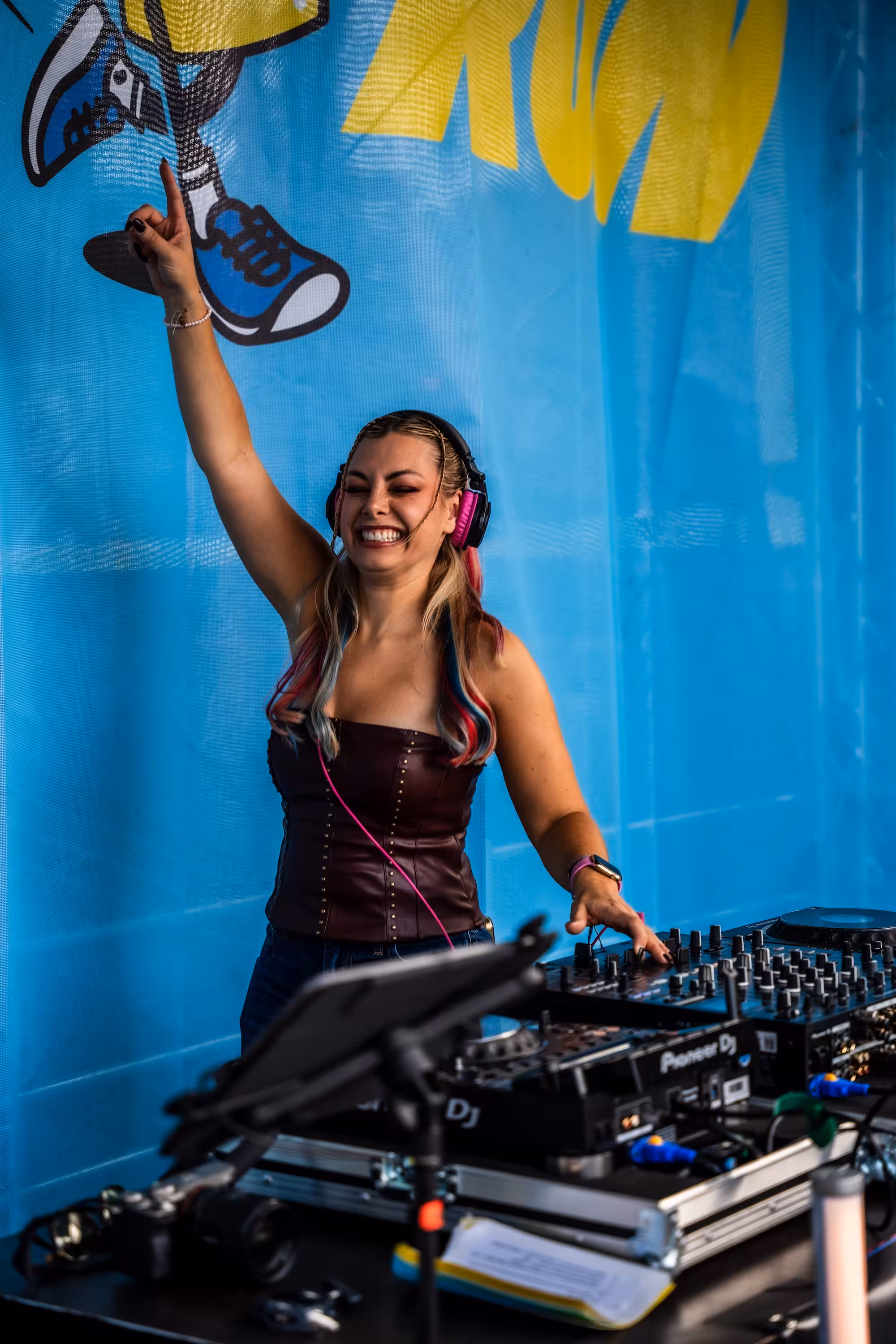 DJ Chany Dakota mit bunten Zöpfen und Kopfhörern hebt hinter ihrem Mischpult den Arm. Sie steht vor einer blauen Wand mit dem HOLY Hotfoot Run Logo.
