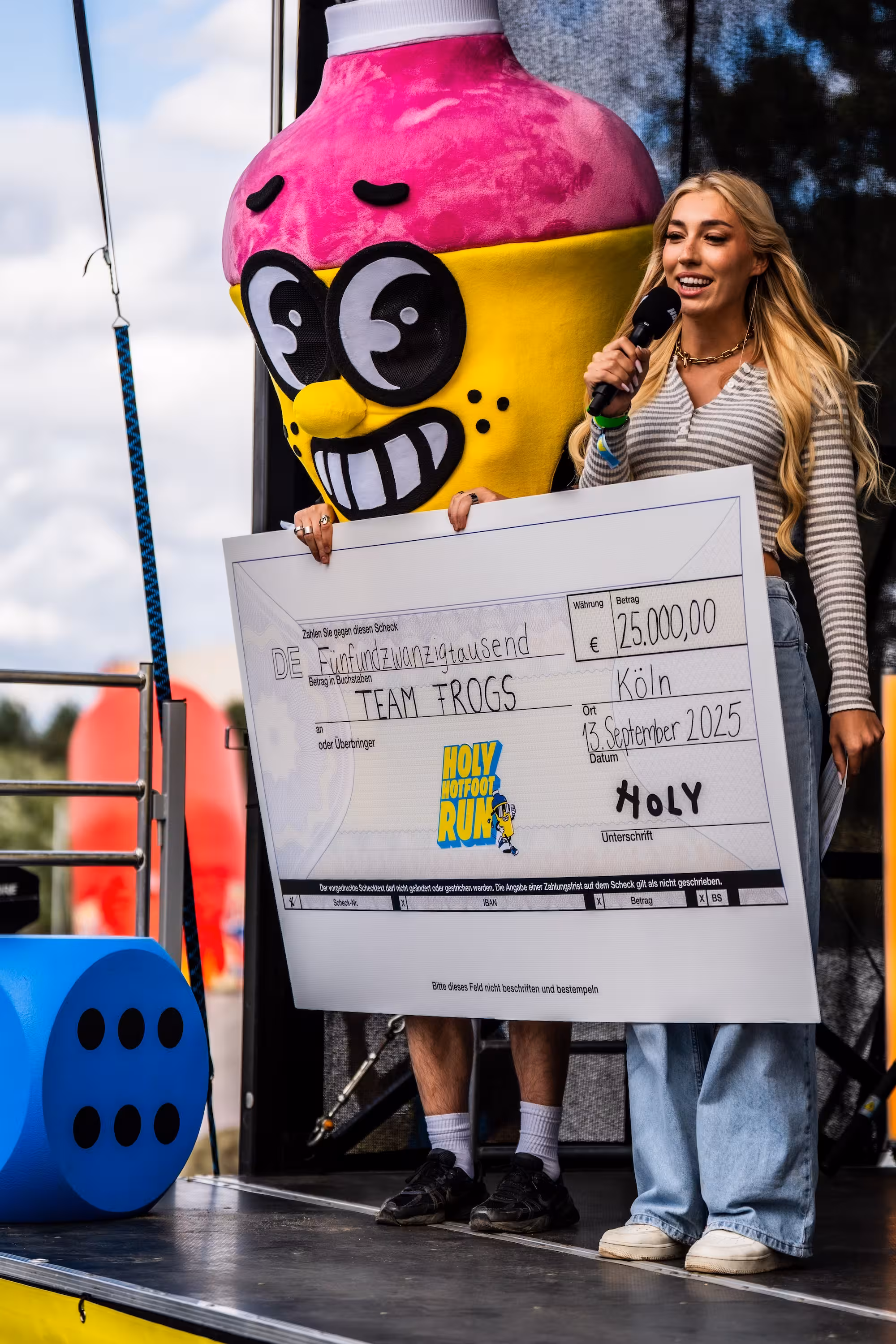 Scheckübergabe auf der Bühne: Eine Moderatorin und das HOLY Maskottchen halten einen großen Scheck über 25.000 Euro für das 'TEAM FROGS' in die Kamera.