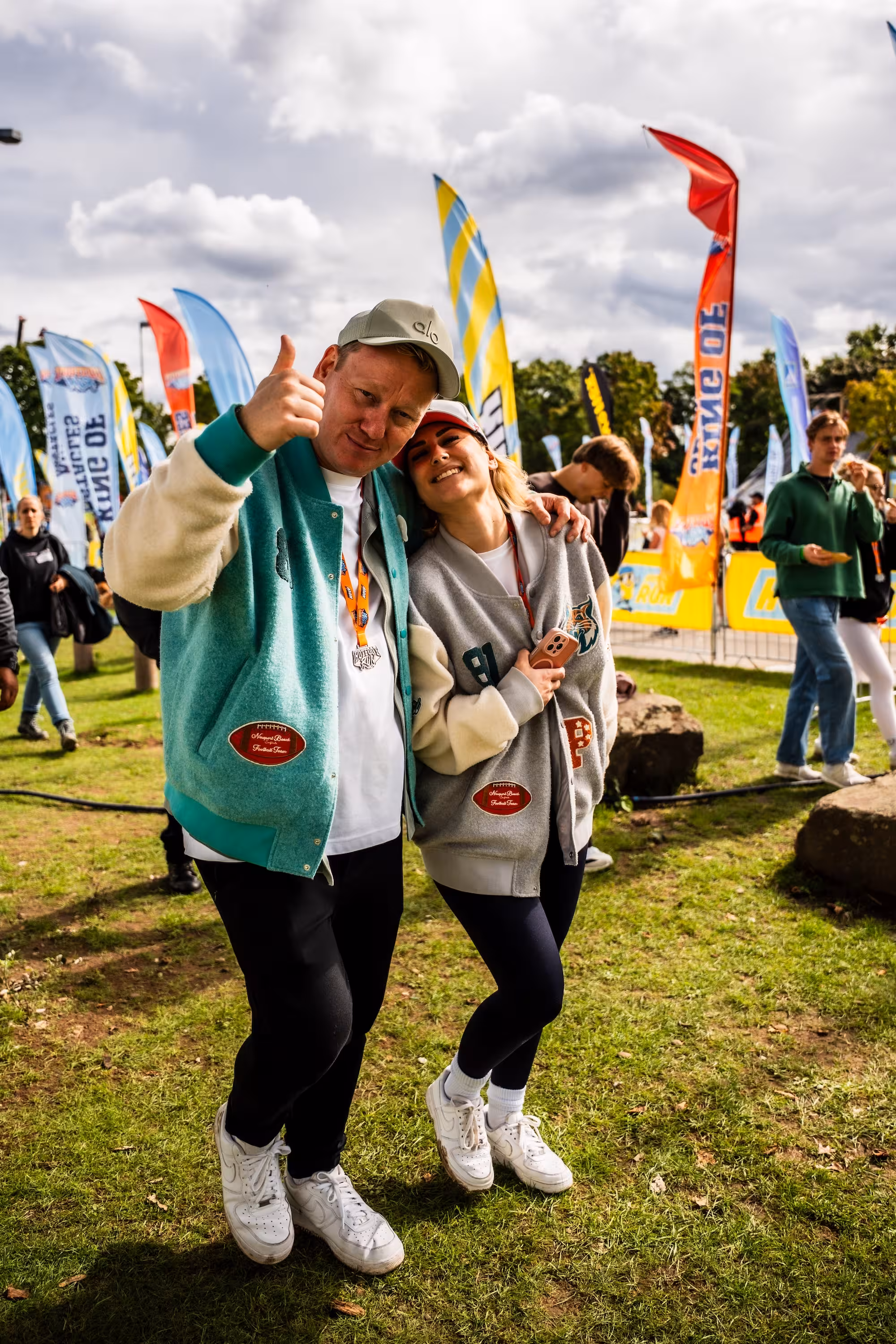 Entertainer Jens 'Knossi' Knossalla zeigt den Daumen hoch, während eine Frau in grauer PARI College-Jacke sich an ihn lehnt. Im Hintergrund wehen bunte Fahnen auf der Wiese.