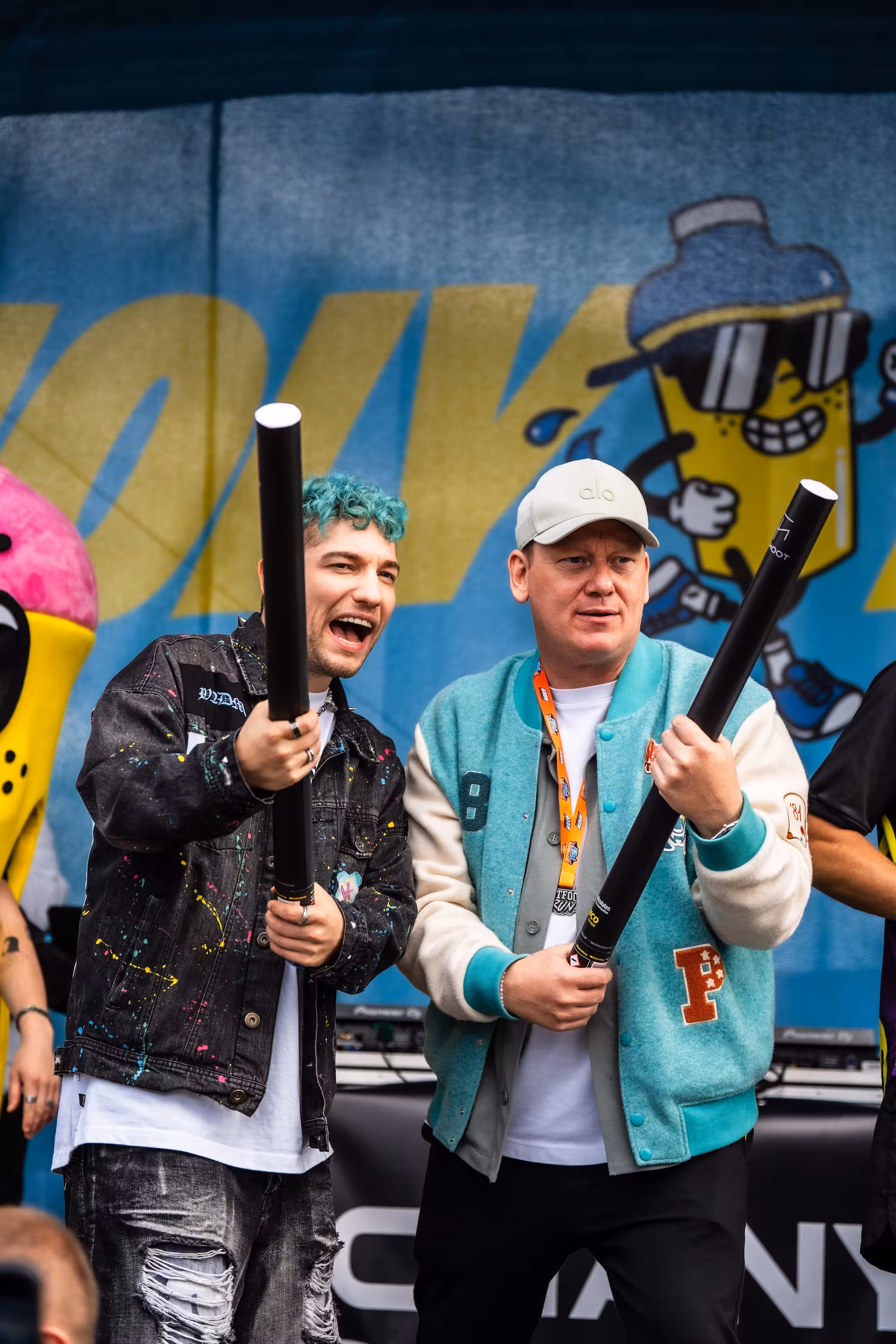 Die Webstars Rezo und Knossi halten große schwarze Konfetti-Kanonen in den Händen. Sie stehen vor einer Bühne mit dem gelben HOLY Maskottchen im Hintergrund.