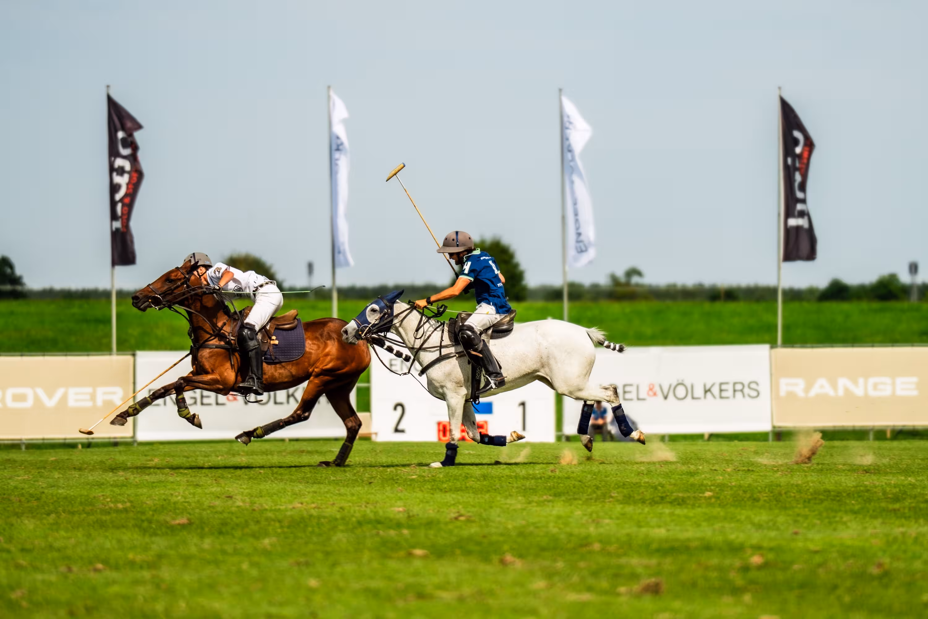 Dynamische Spielszene: Ein Spieler auf einem Schimmel reitet schnell an einem Gegner auf einem braunen Pferd vorbei. Im Hintergrund wehen Sponsorenfahnen im Wind.