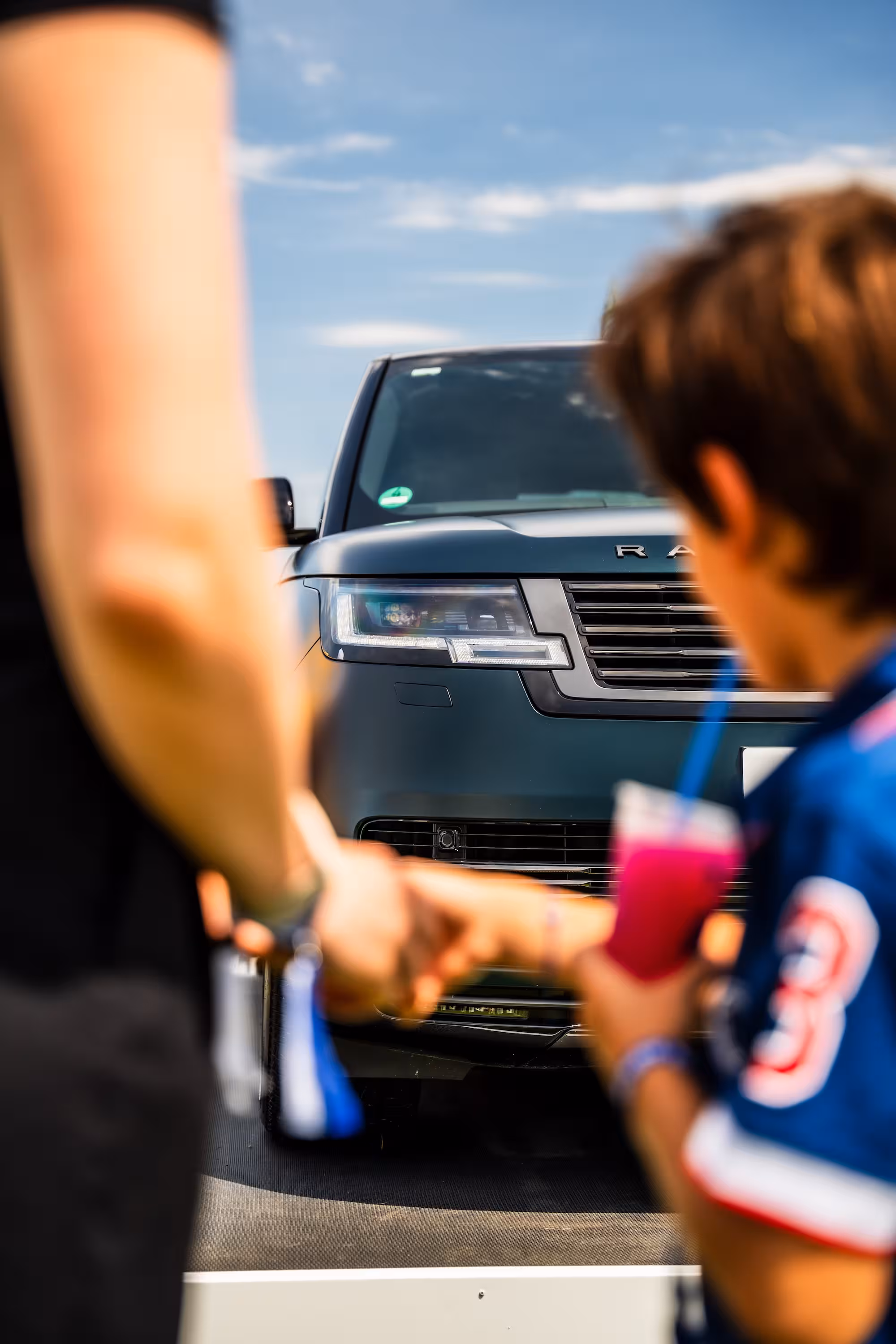 Unscharf im Vordergrund: Ein Kind mit blauem Trikot und eine Erwachsene halten einen Schlüssel in den Händen. Im Fokus dahinter steht die Front eines dunkelgrünen Range Rovers auf einem Eventgelände.