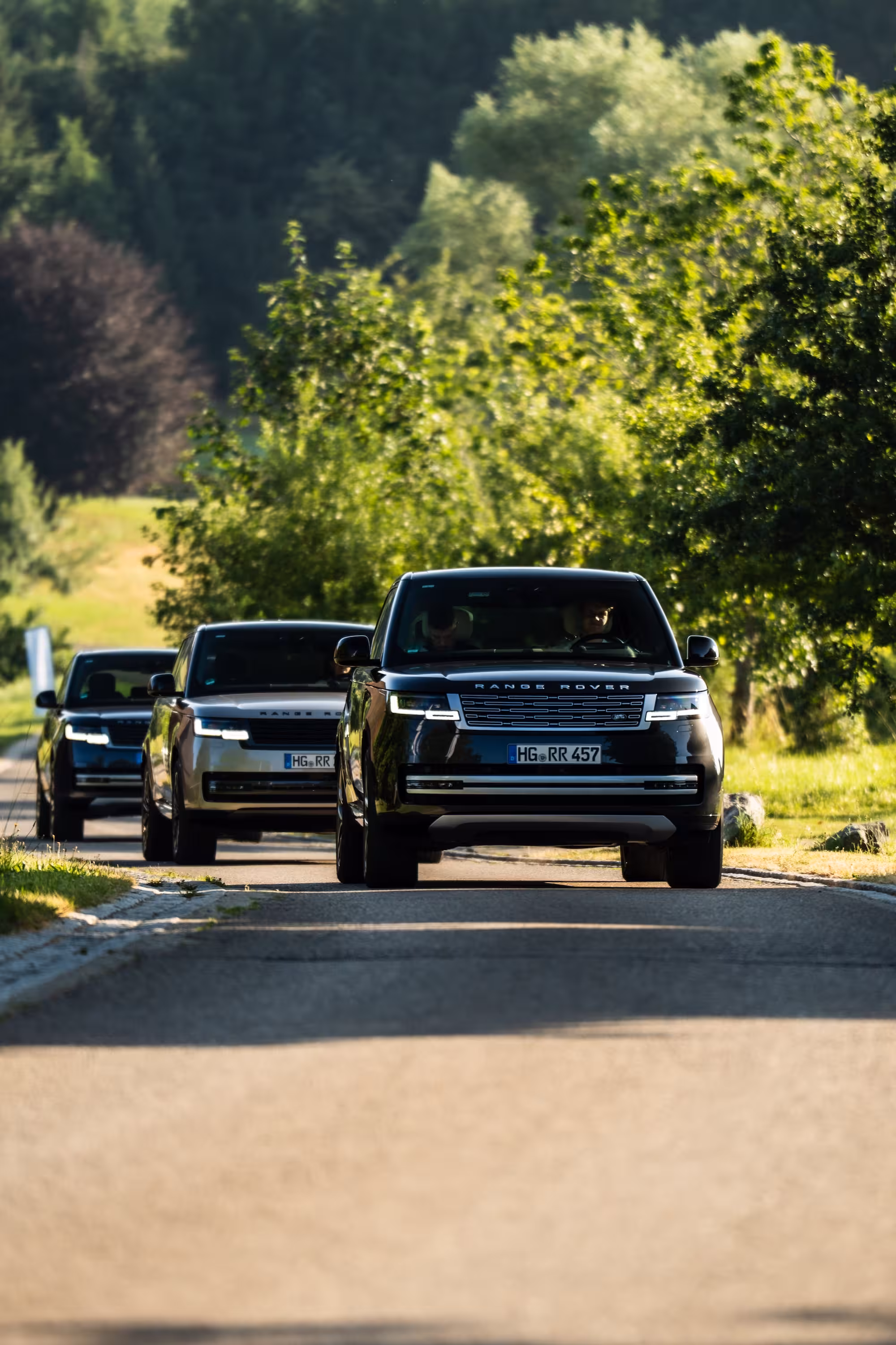 Konvoi aus drei Range Rover Fahrzeugen fährt hintereinander auf einer sonnigen Landstraße, gesäumt von grünen Bäumen.