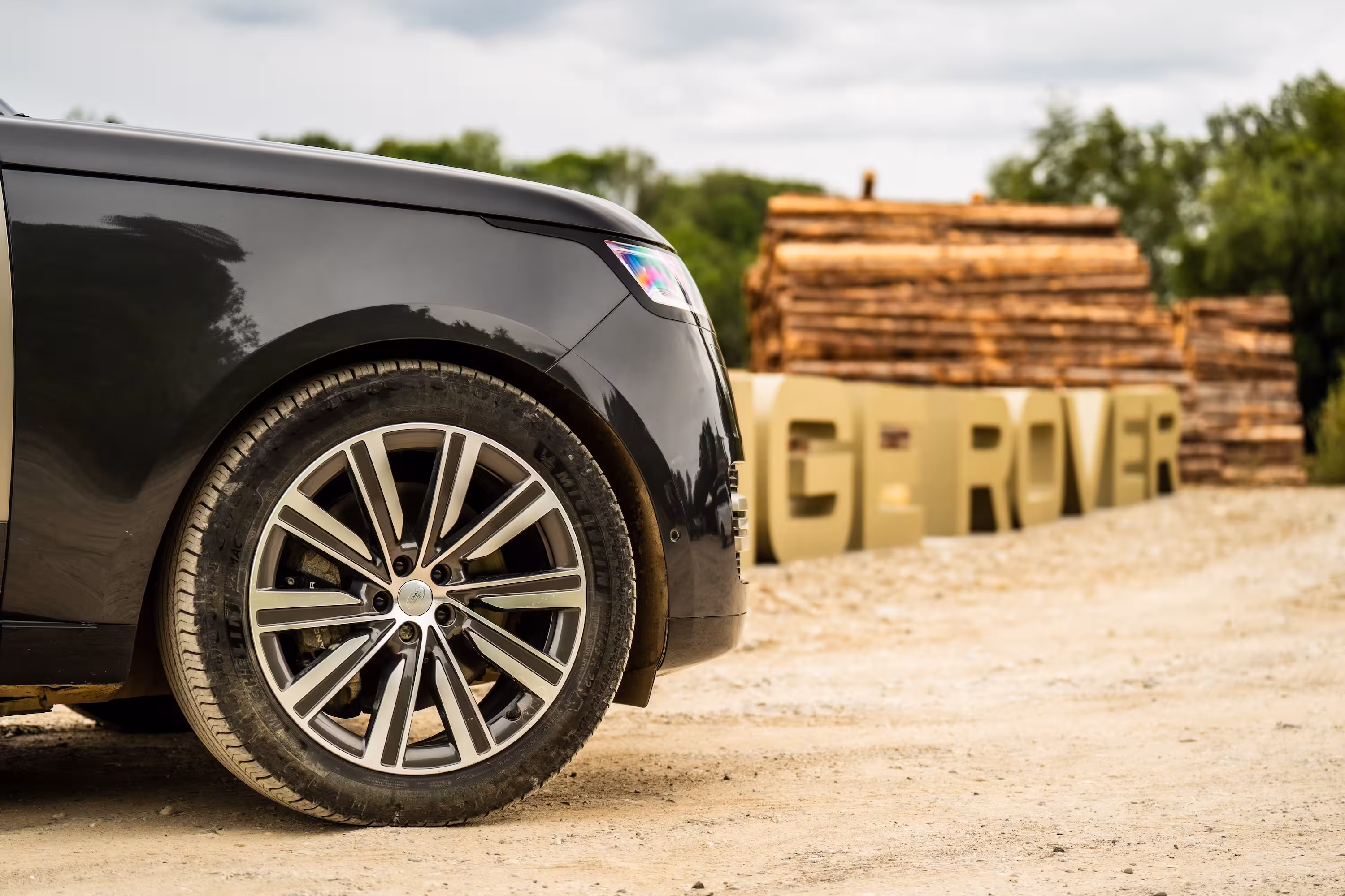 Nahaufnahme des Vorderrads und der Felge eines schwarzen Range Rovers. Im unscharfen Hintergrund stehen große, goldene Buchstaben, die den Schriftzug 'RANGE ROVER' bilden.
