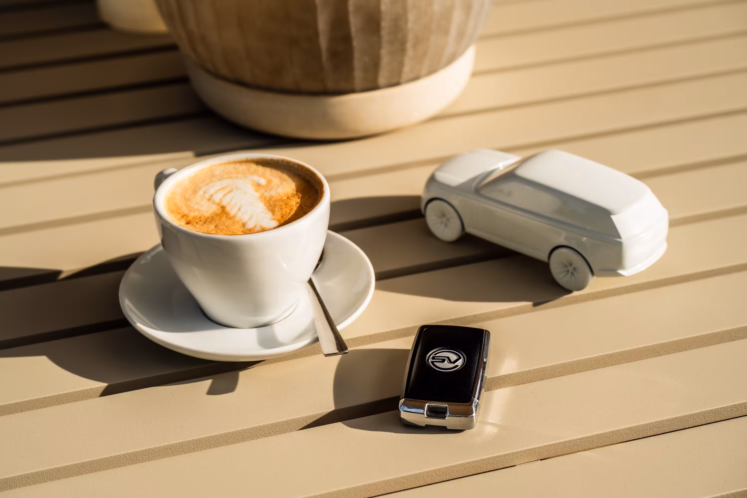 Detailaufnahme auf einem Tisch: Eine Tasse Cappuccino mit Latte Art, ein stilisiertes weißes Keramik-Modellauto und ein Range Rover SV Autoschlüssel liegen im Sonnenlicht.