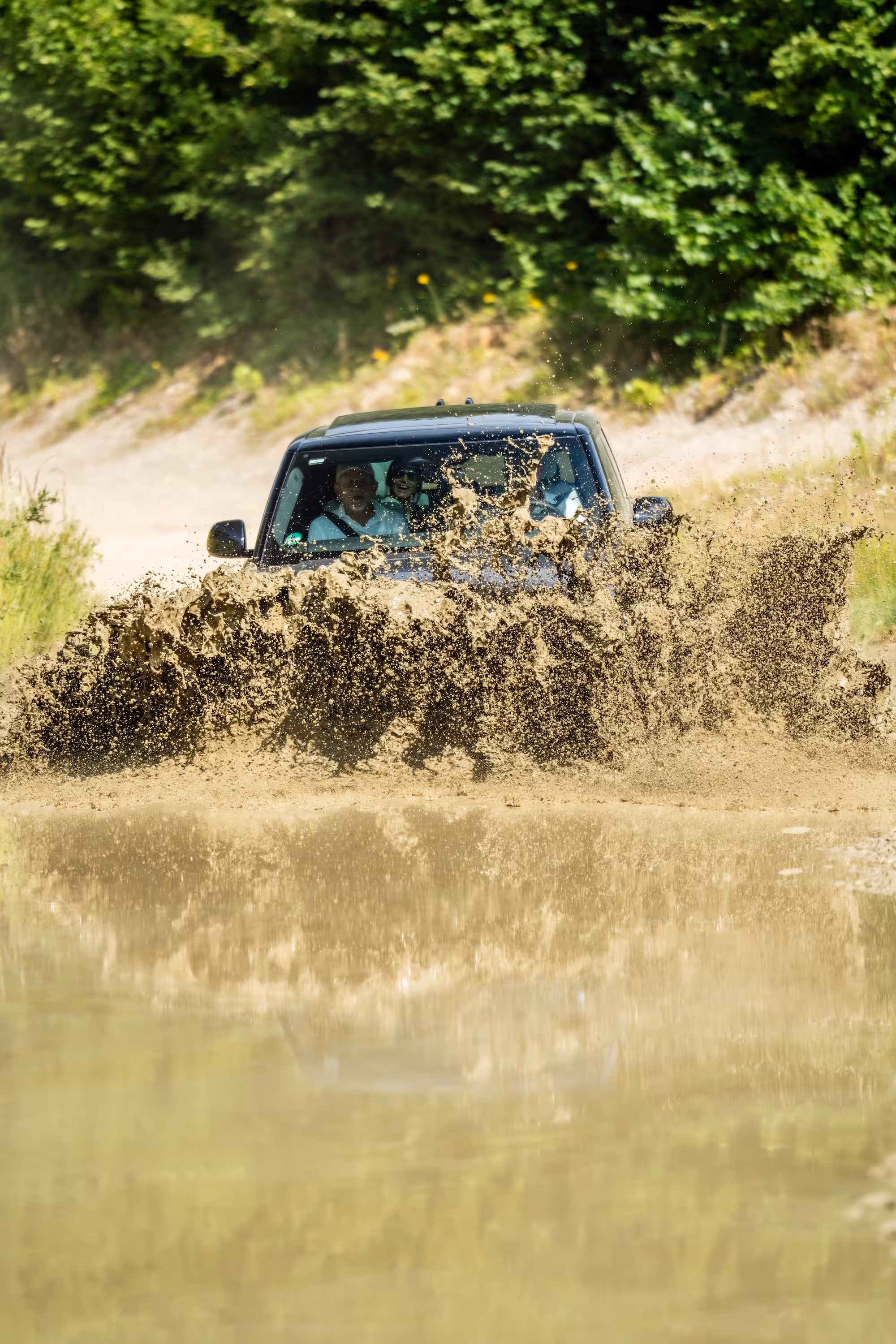 Actionreiche Offroad-Fahrt: Ein schwarzer Range Rover fährt durch ein tiefes Wasserloch. Eine riesige Fontäne aus braunem Wasser spritzt vor dem Fahrzeug hoch.