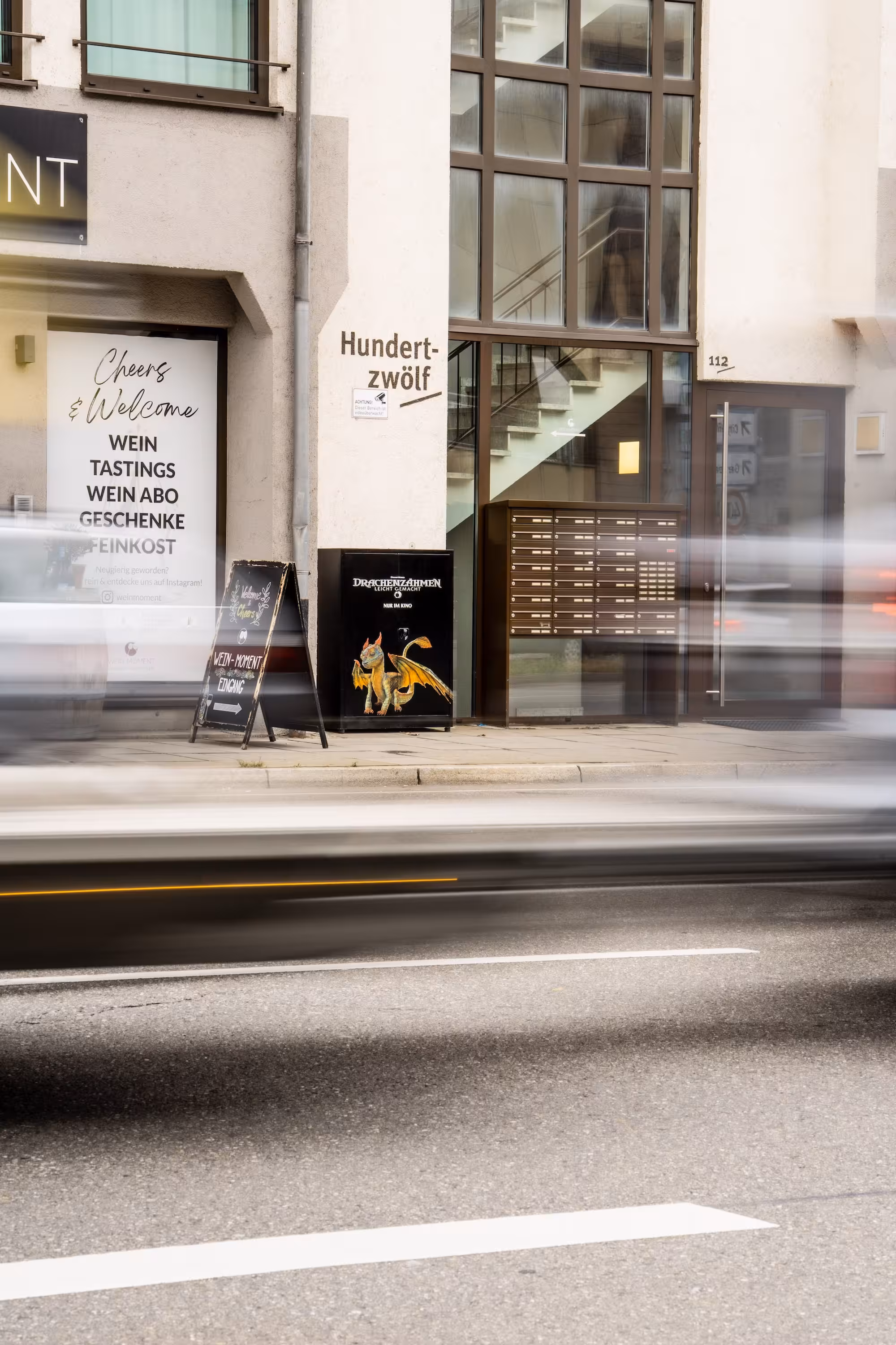 schwarzer Stromkasten mit Drachen-Motiv steht an einer Straße in Stuttgart vor einem Gebäude. Vorbeifahrende Autos sind durch Bewegungsunschärfe verwischt. Teil der 'Great Dragon Hunt' Kampagne von Die Mondpiloten.