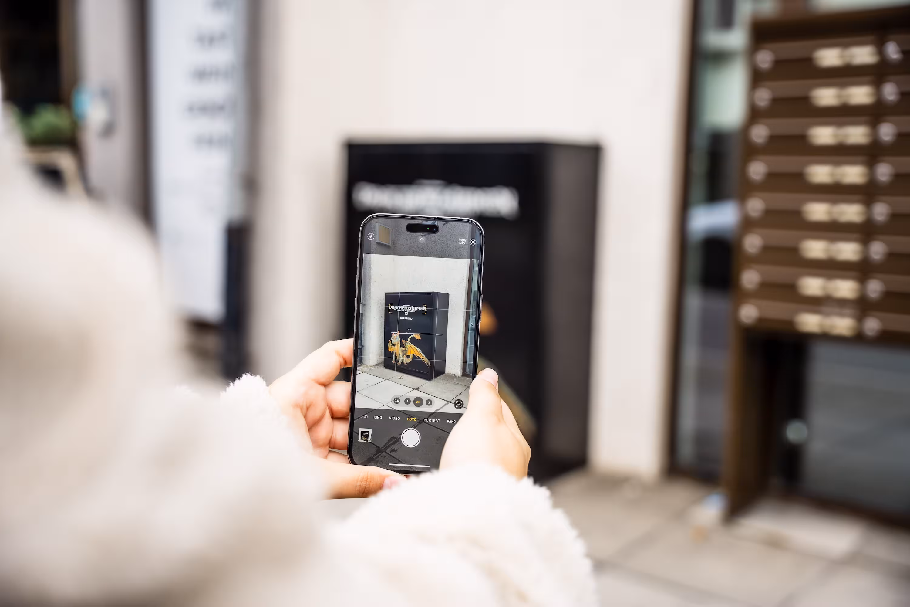 Blick auf ein Smartphone-Display, das gerade ein Foto des Drachen-Stromkastens macht. Die Hand hält das Handy hochkant, im Hintergrund ist der schwarze Kasten unscharf zu sehen.