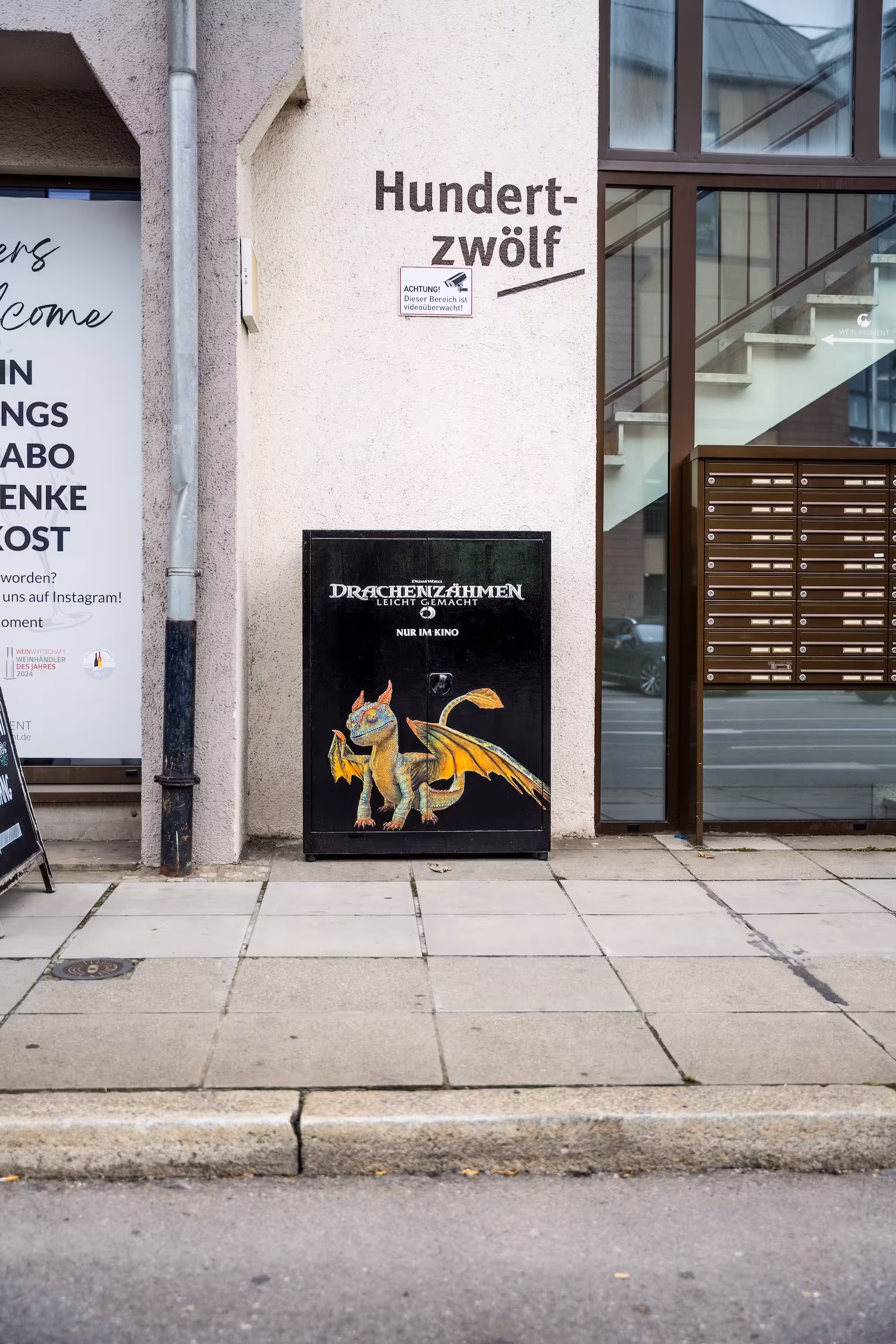 Frontalansicht des schwarzen Stromkastens vor einer Hauswand. Darauf das weiße Filmlogo 'Drachenzähmen leicht gemacht' und darunter eine farbenfrohe Illustration eines sitzenden Drachen.