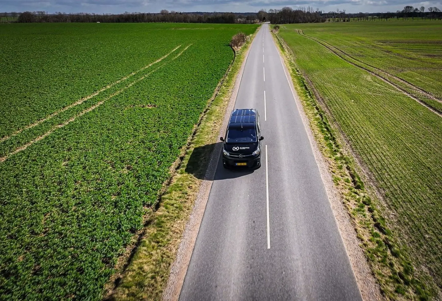 Black van with a company logo driving on a narrow rural road surrounded by green fields under a cloudy sky.