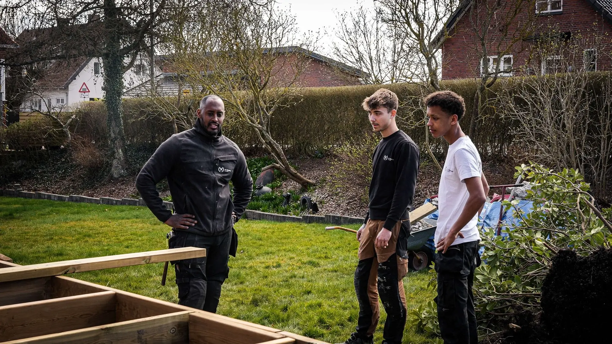 Three men standing on a lawn near wooden construction materials, appearing to discuss a building project.