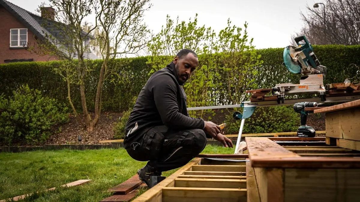 Tømrer monterer terrassefundament i have med kap- og geringssav i baggrunden
