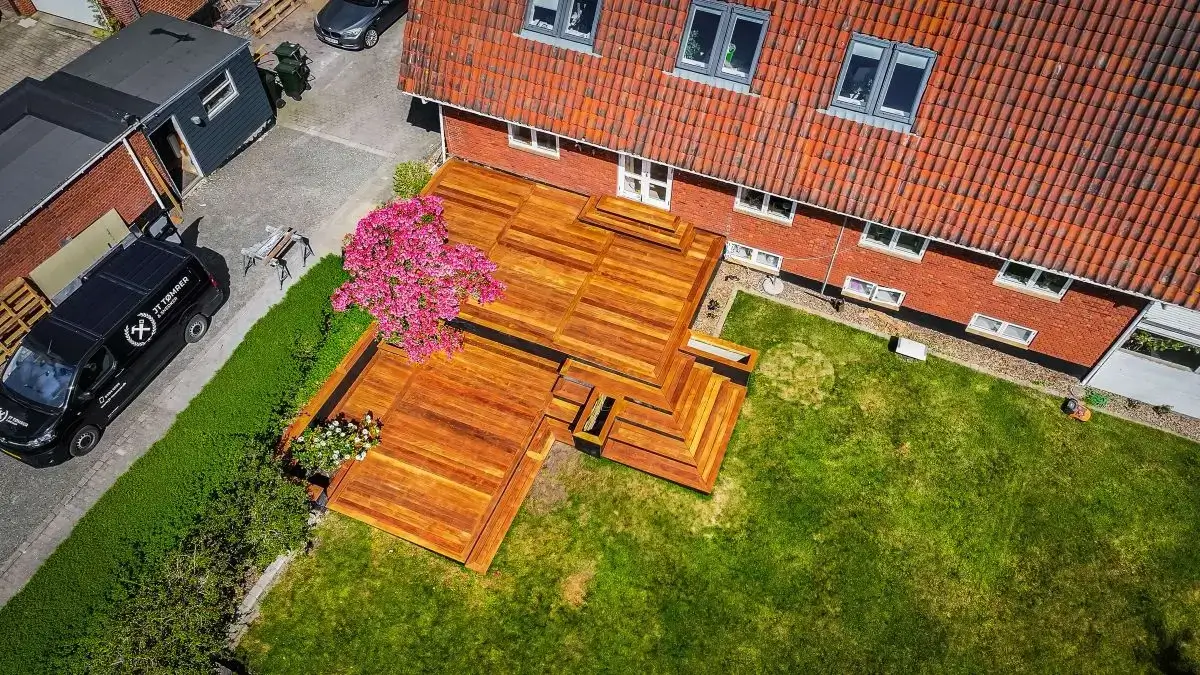 Stor træterrasse med trapper og blomsterbed set fra oven foran murstenshus.