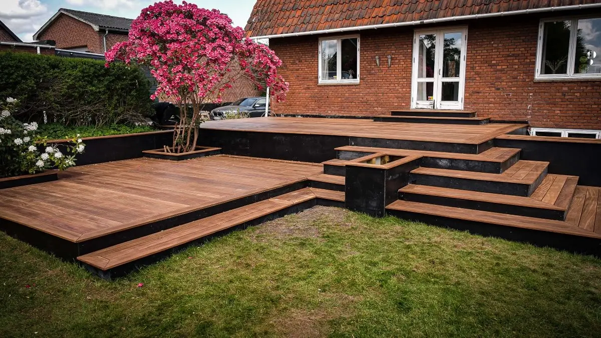Stor færdigbygget træterrasse med trapper og blomsterbed foran villa.