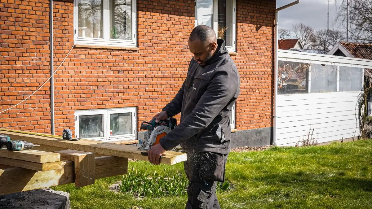 Tømrer saver træbjælke til terrasse med rundsav foran murstenshus
