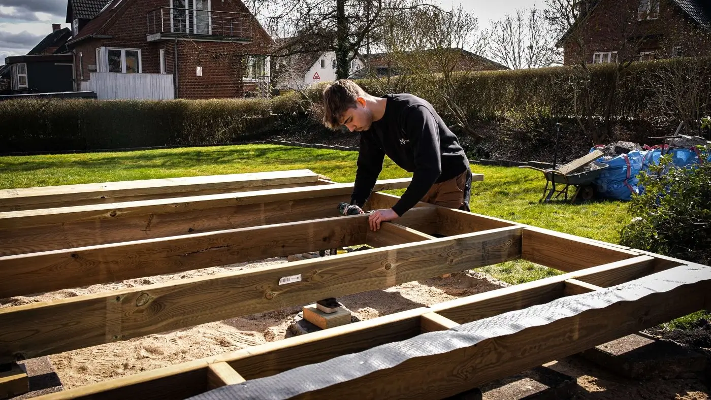 Tømrer monterer træbjælker til fundamentet for ny terrasse i have.