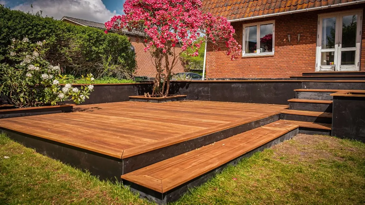 Nybygget træterrasse med blomsterbed og trappe foran murstenshus.