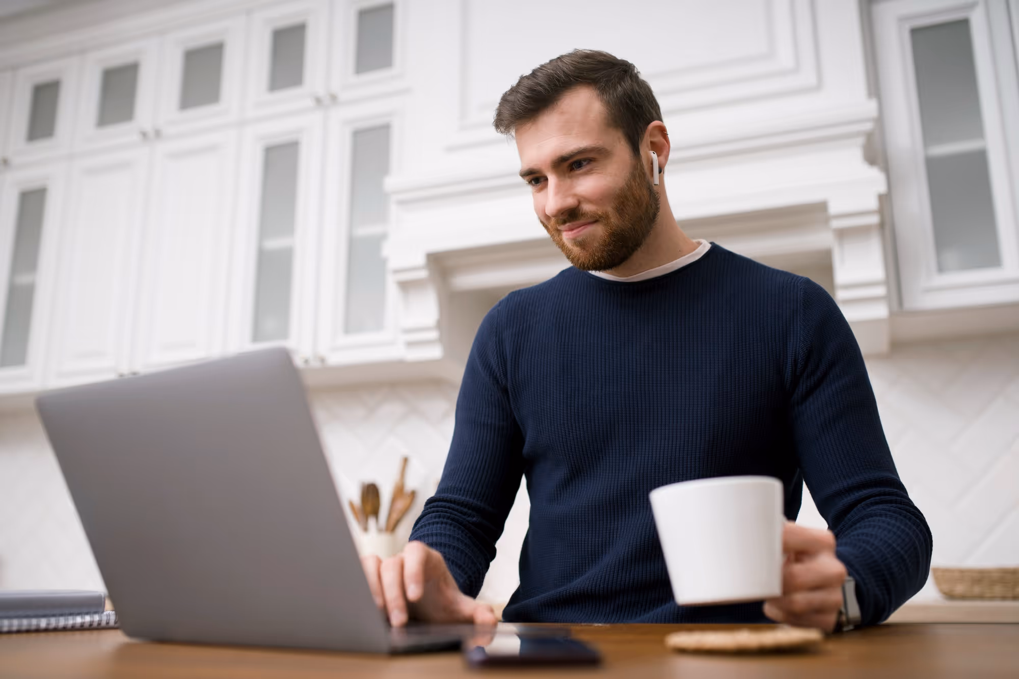 Man in a navy sweater wearing wireless earbuds, working on a laptop while holding a white mug in a bright kitchen.