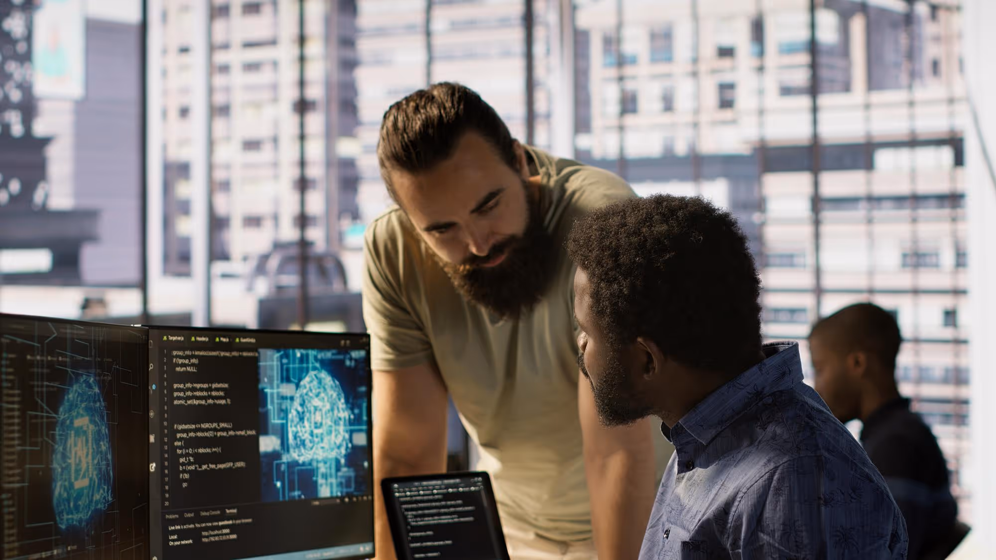 Two men collaborating and looking at coding and a digital brain image on computer monitors in a modern office.