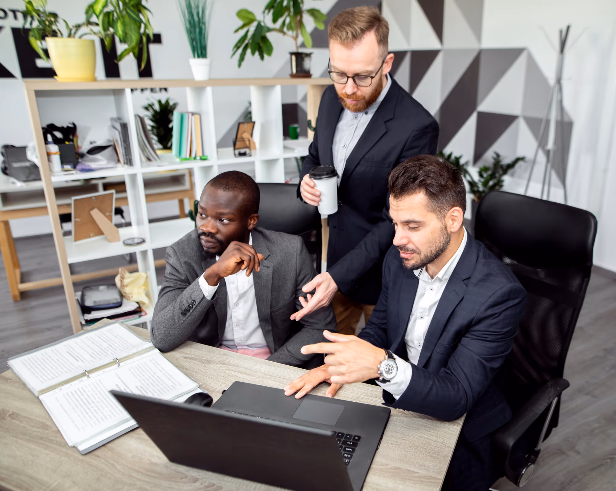 Three businessmen in suits discuss work at a desk with a laptop and open binder in a modern office.