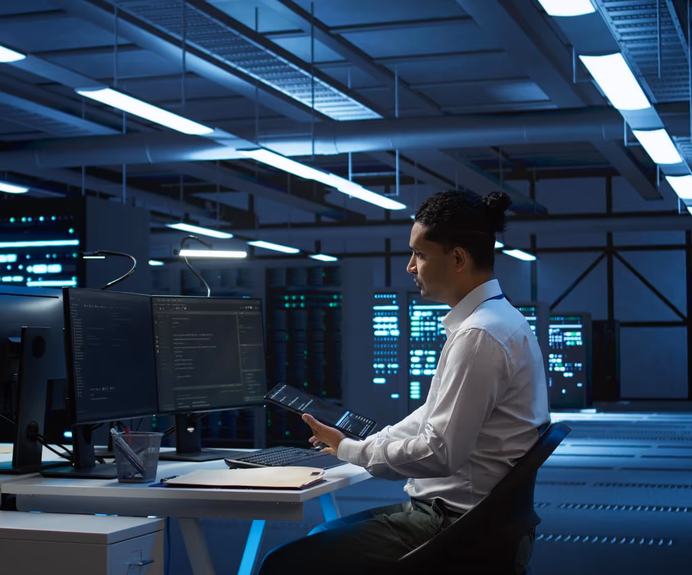 Man in a data center working on a tablet with dual monitors displaying code in a dimly lit server room.