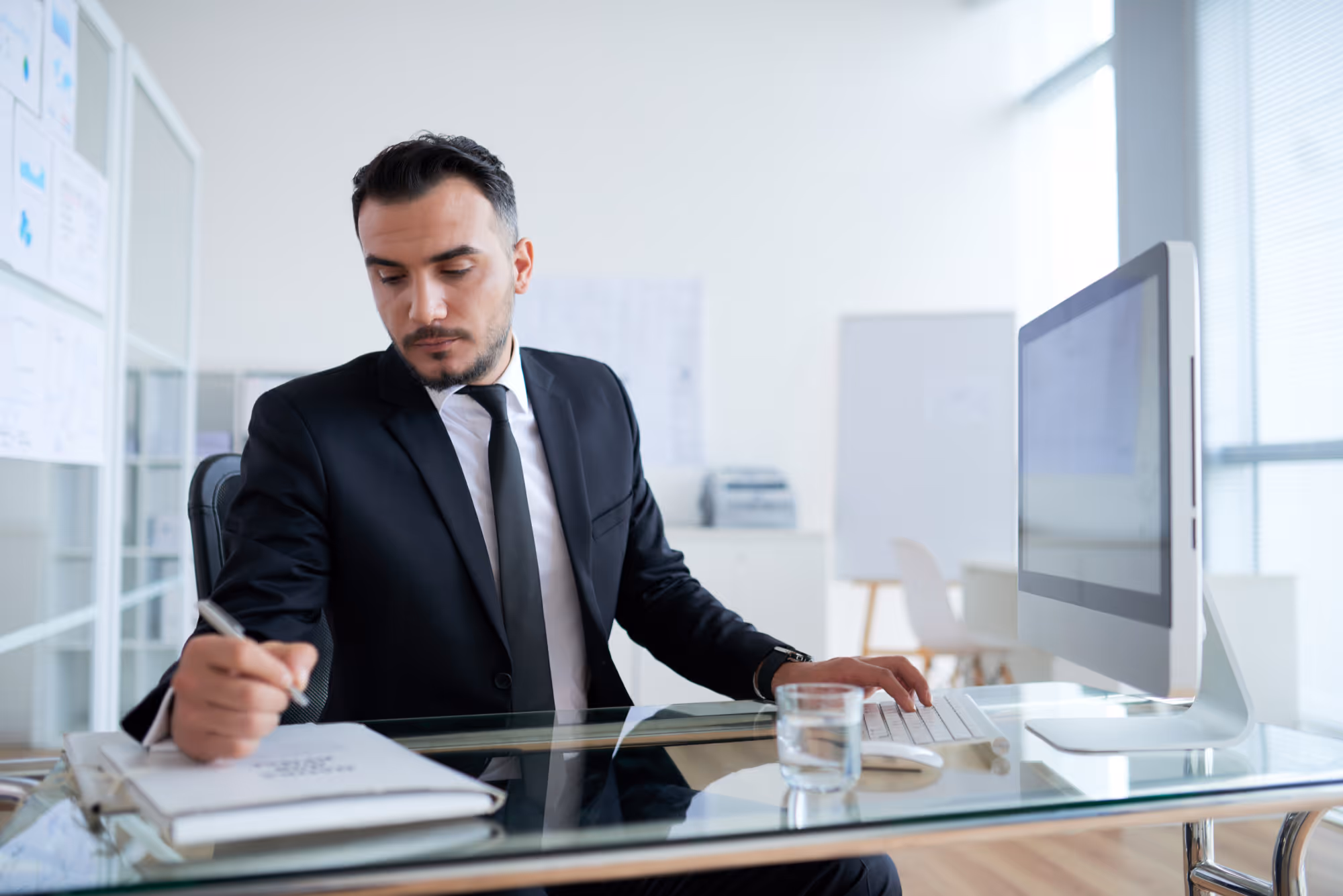 Man in a suit writing in a notebook while sitting at a glass desk with a computer and a glass of water.