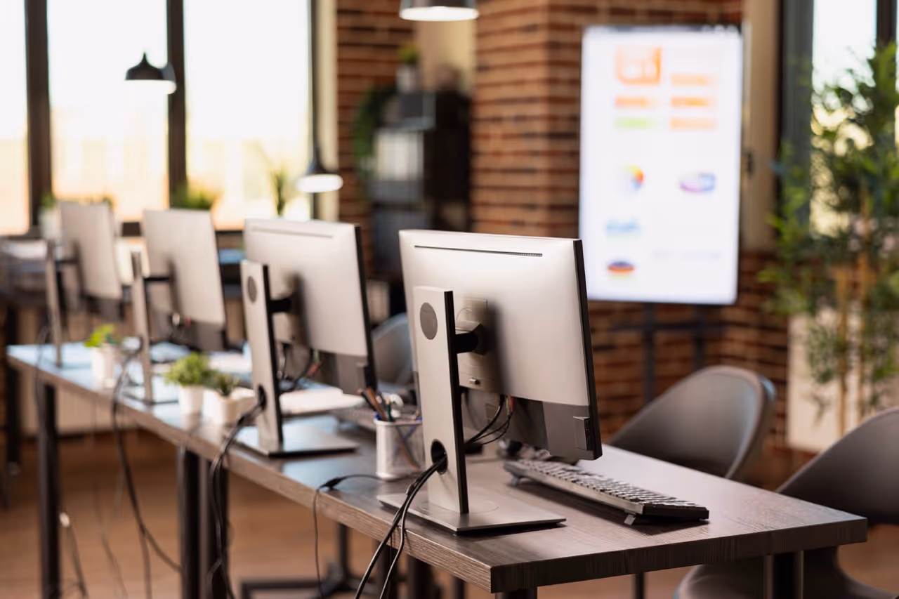 Row of modern computer monitors and keyboards on a long wooden desk in a bright office with brick walls and large windows.
