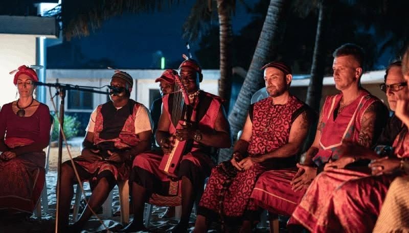 Bwiti practitioners and retreat participants seated together at night in Gabon, one playing a ngombi harp.