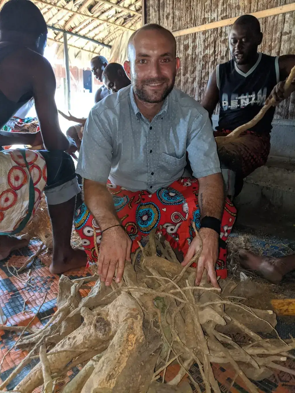 Ryan 'Ghenigho' Rich kneels smiling behind a large pile of Iboga roots, with Bwiti practitioners in a traditional thatched-roof hut during Iboga preparation.