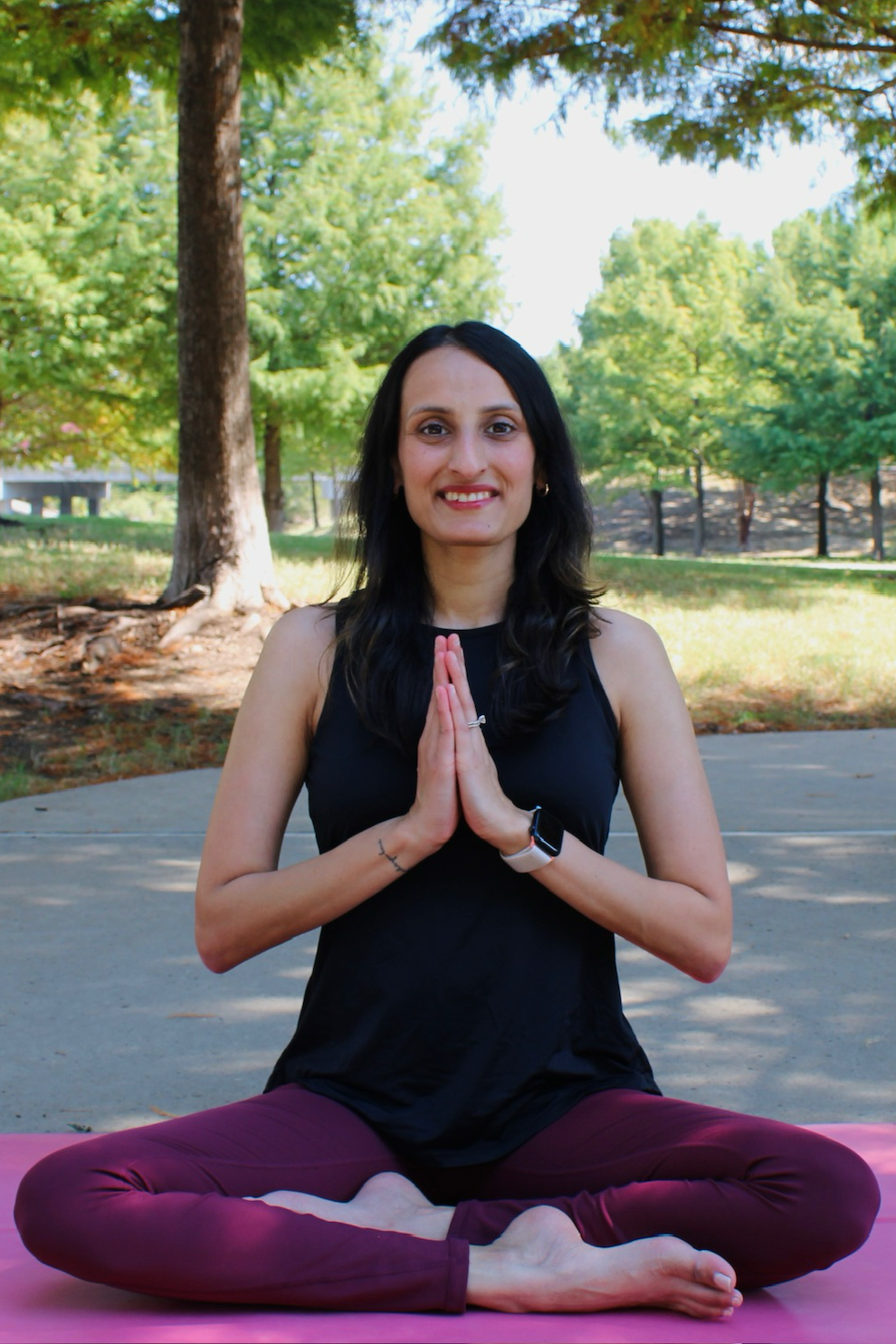 Woman sitting cross-legged on a pink yoga mat outdoors, with hands pressed together in a prayer pose, smiling at the camera.