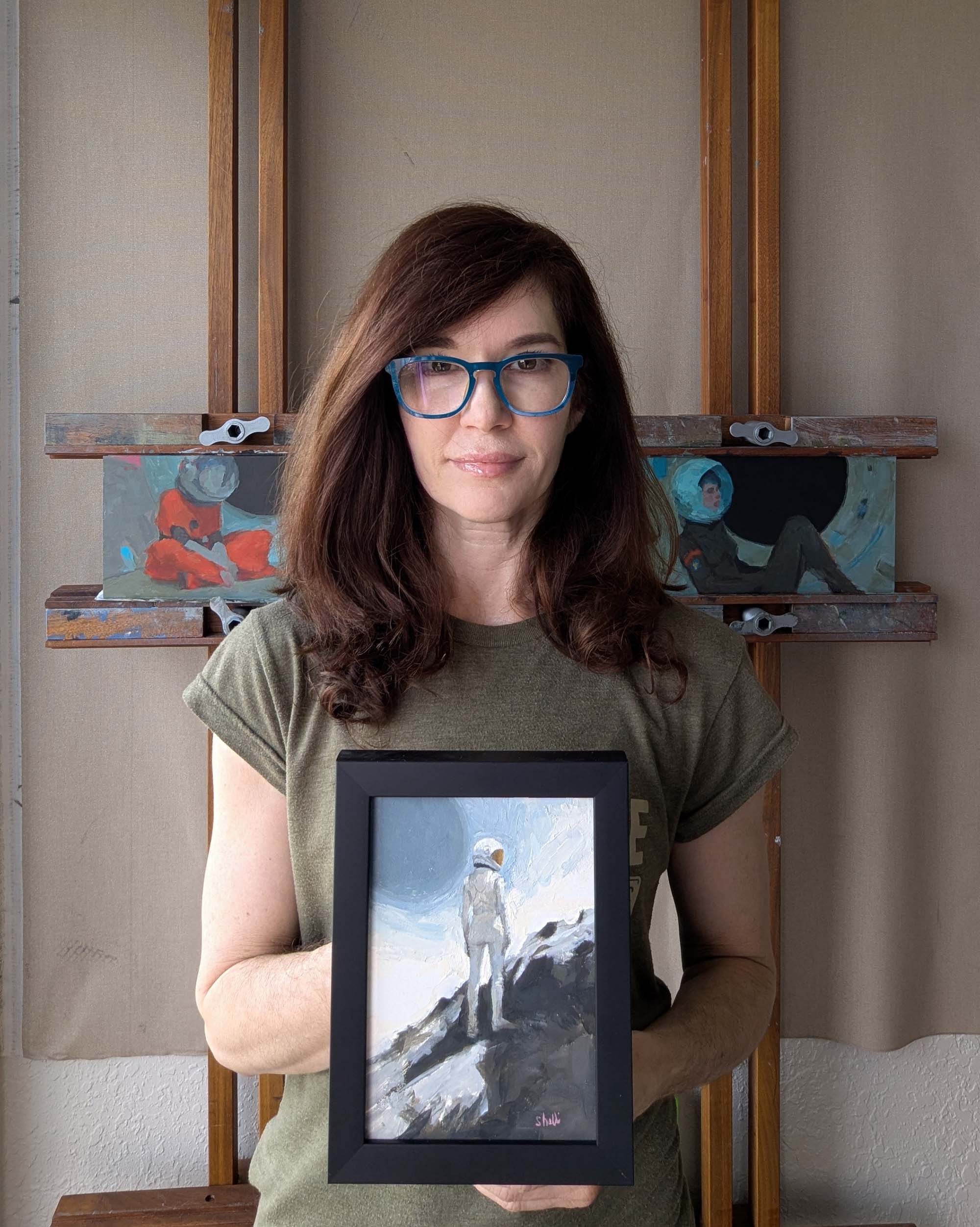 artist holding oil painting of a space explorer standing on a hill looking Eastward. A blue celestial body in the sky at top left.