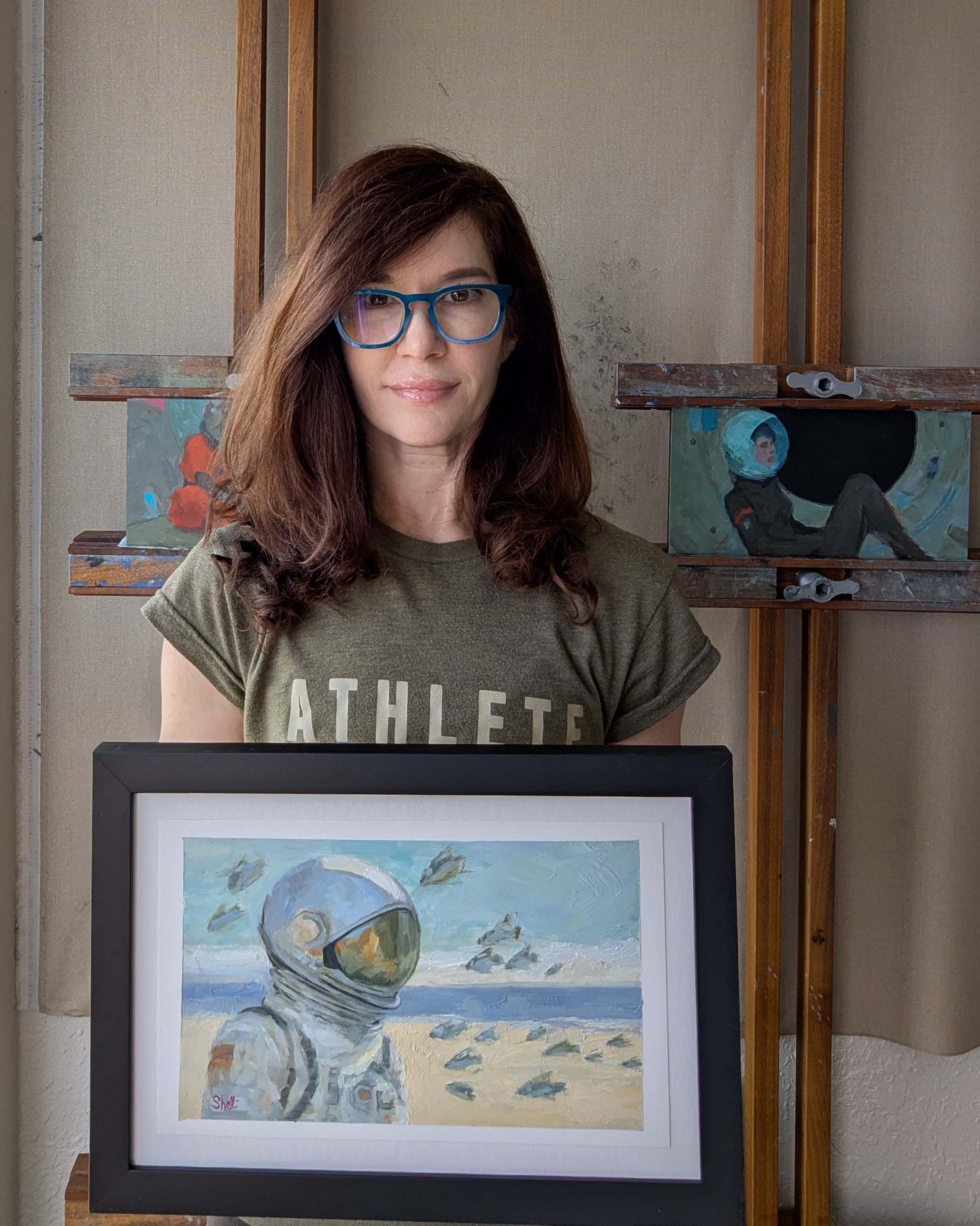 artist holding painting of a space explorer on the beach facing East with fish swimming in the air around her.